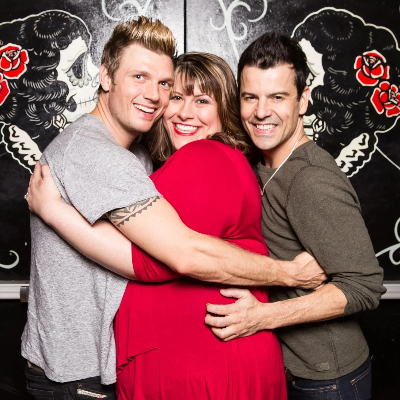 Three people hugging and smiling in front of a black wall with decorative artwork, including two stylized faces with roses.
