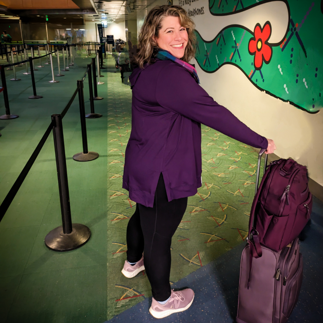 A woman with curly hair smiling at the airport, standing in line with purple luggage, wearing a purple jacket, black leggings, and pink sneakers, near a colorful mural with a flower on the wall.