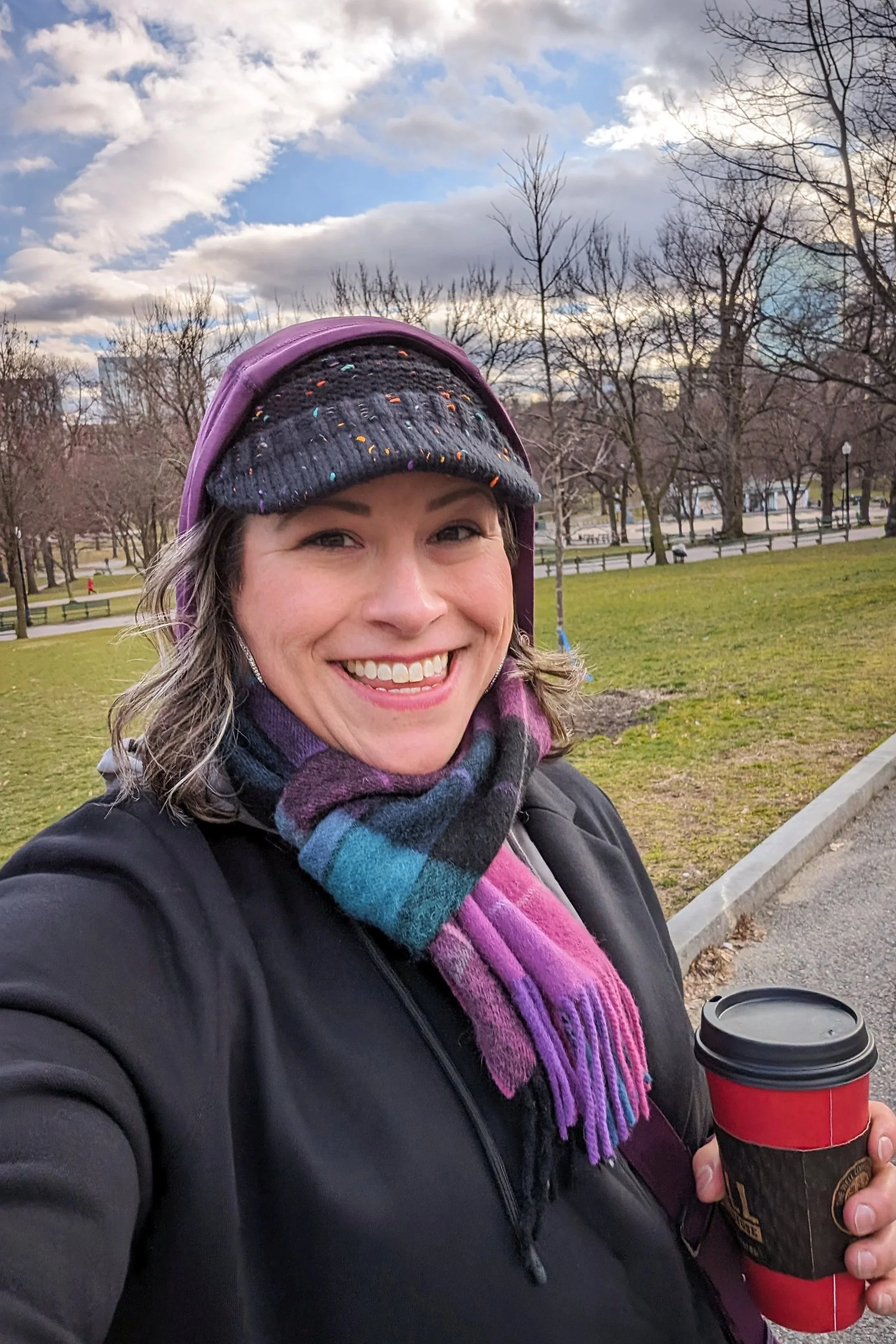A woman outside in a park on a cloudy day, smiling and holding a coffee cup, wearing a black jacket, colorful scarf, hat, and purple backpack.