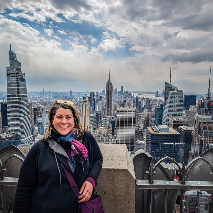 A smiling woman standing on a rooftop observation deck with New York City skyline in the background, including the Empire State Building and skyscrapers under a cloudy sky.