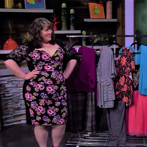 A woman in a floral dress standing in a clothing store or boutique, smiling and looking at a rack of various colorful clothes.