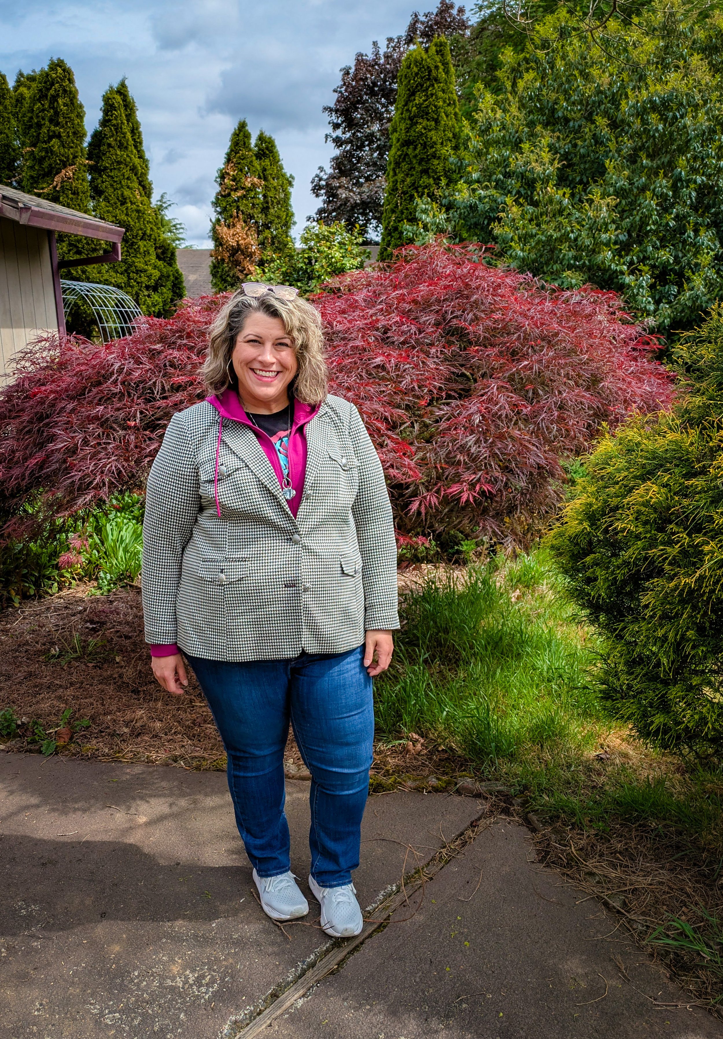 A woman smiling outdoors in front of a red leafed shrub, with trees and a cloudy sky in the background.