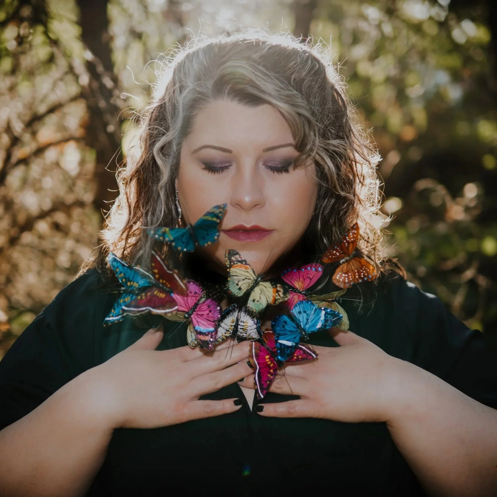 A woman with curly hair and makeup, standing outdoors with blurred trees in the background, holding a cluster of colorful butterflies near her collarbone with her hands.