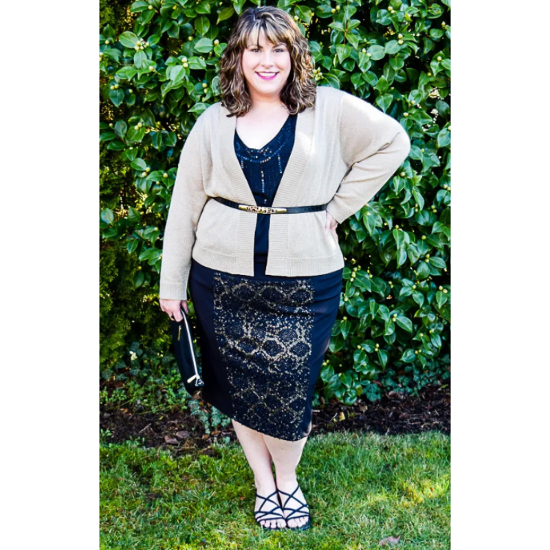 A woman standing outdoors in front of green foliage, wearing a beige cardigan, black shirt, patterned black and gold skirt, and black strappy heels.
