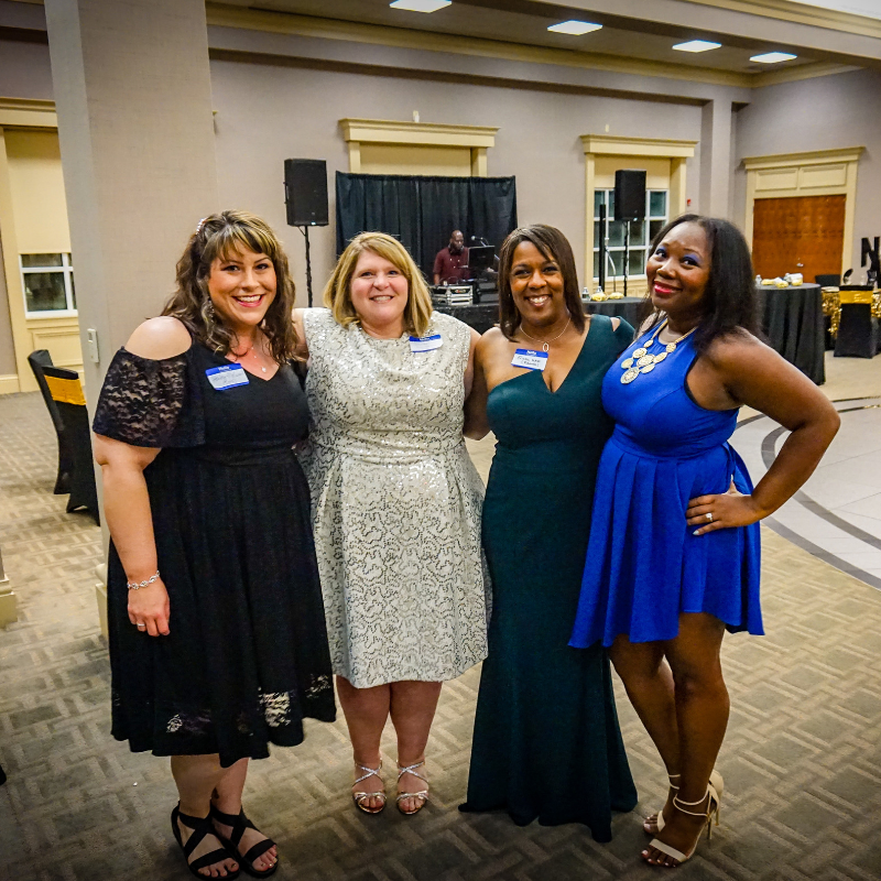 Four women dressed in formal attire standing together at an indoor event with a man in the background near a DJ booth.