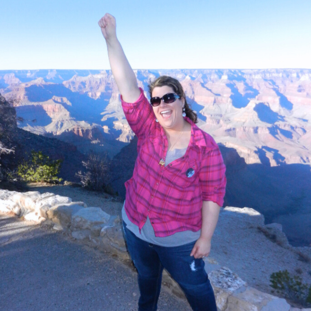 A woman in sunglasses, pink plaid shirt, and jeans standing at the edge of the Grand Canyon, raising her right fist in the air and smiling.