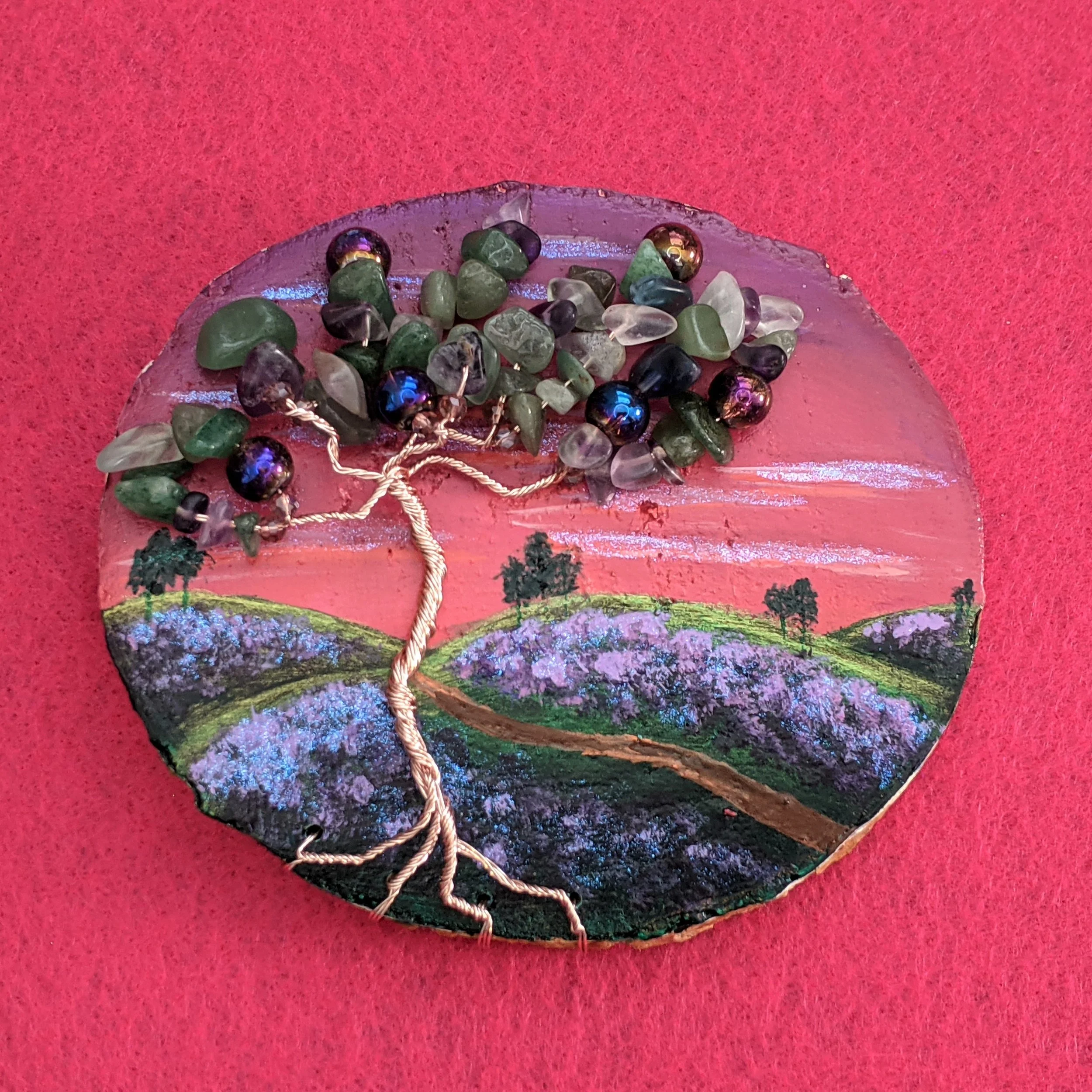 Decorative art piece featuring a painted landscape with hills and trees, topped with a wire tree sculpture adorned with various green, purple, and black beads representing leaves.