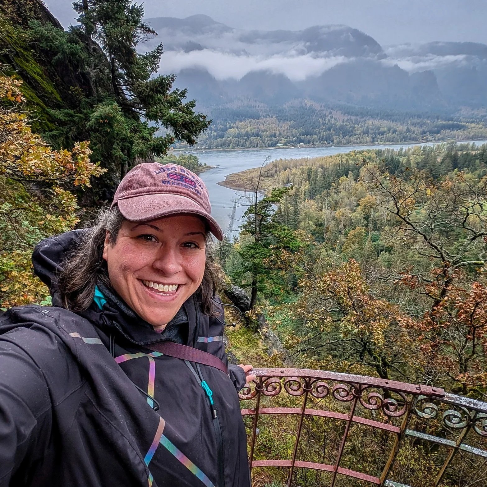 A woman smiling in outdoor hiking gear taking a selfie on a railing overlooking a river and forested mountains.