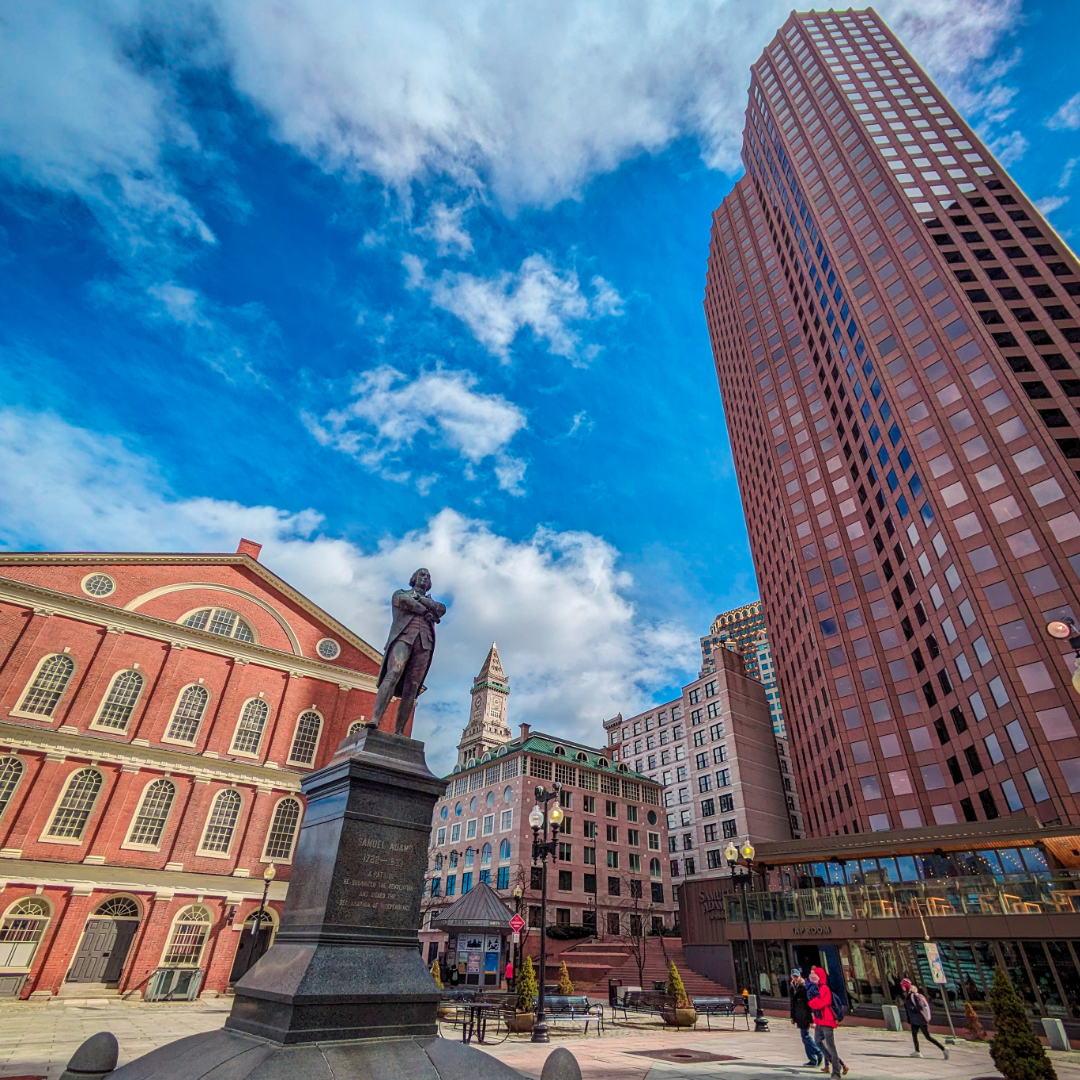 Cityscape with historic and modern buildings, a statue of a man, and a few pedestrians under a partly cloudy sky.
