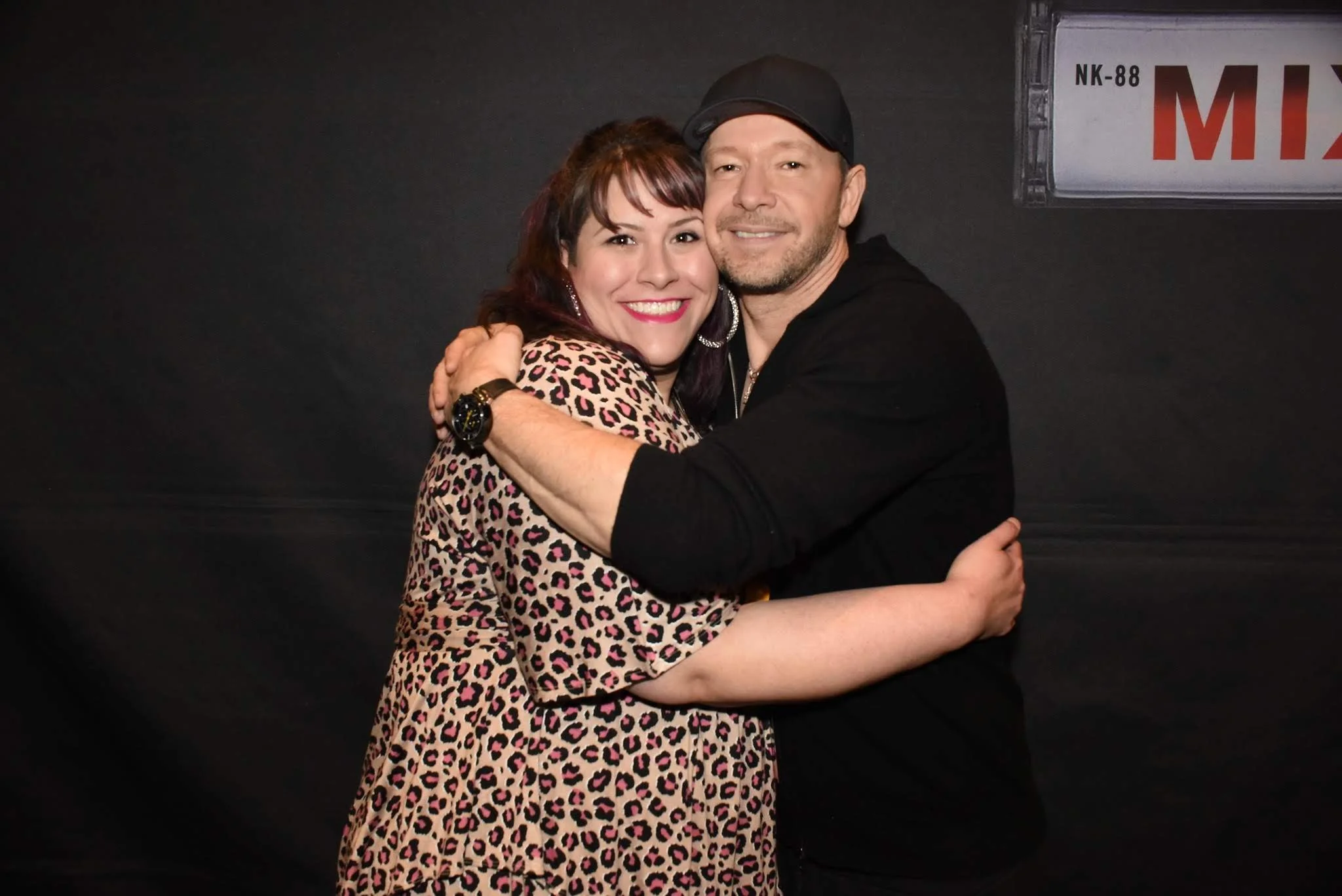 A smiling couple hugging in front of a dark background, with a sign in the top right corner that reads 'MIX'.