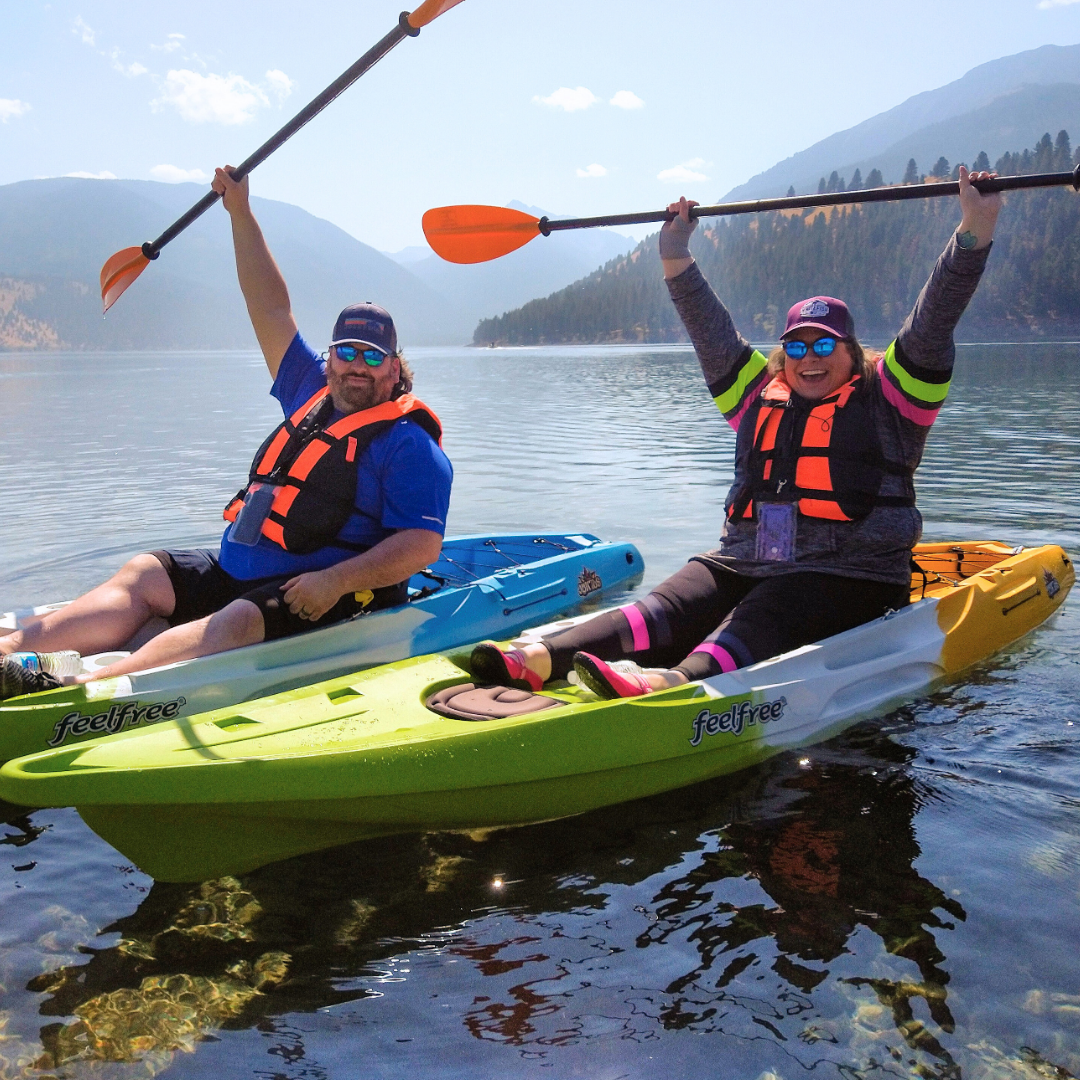 Two people kayaking on a lake surrounded by mountains, wearing life jackets and sunglasses, holding paddles raised in the air and smiling.