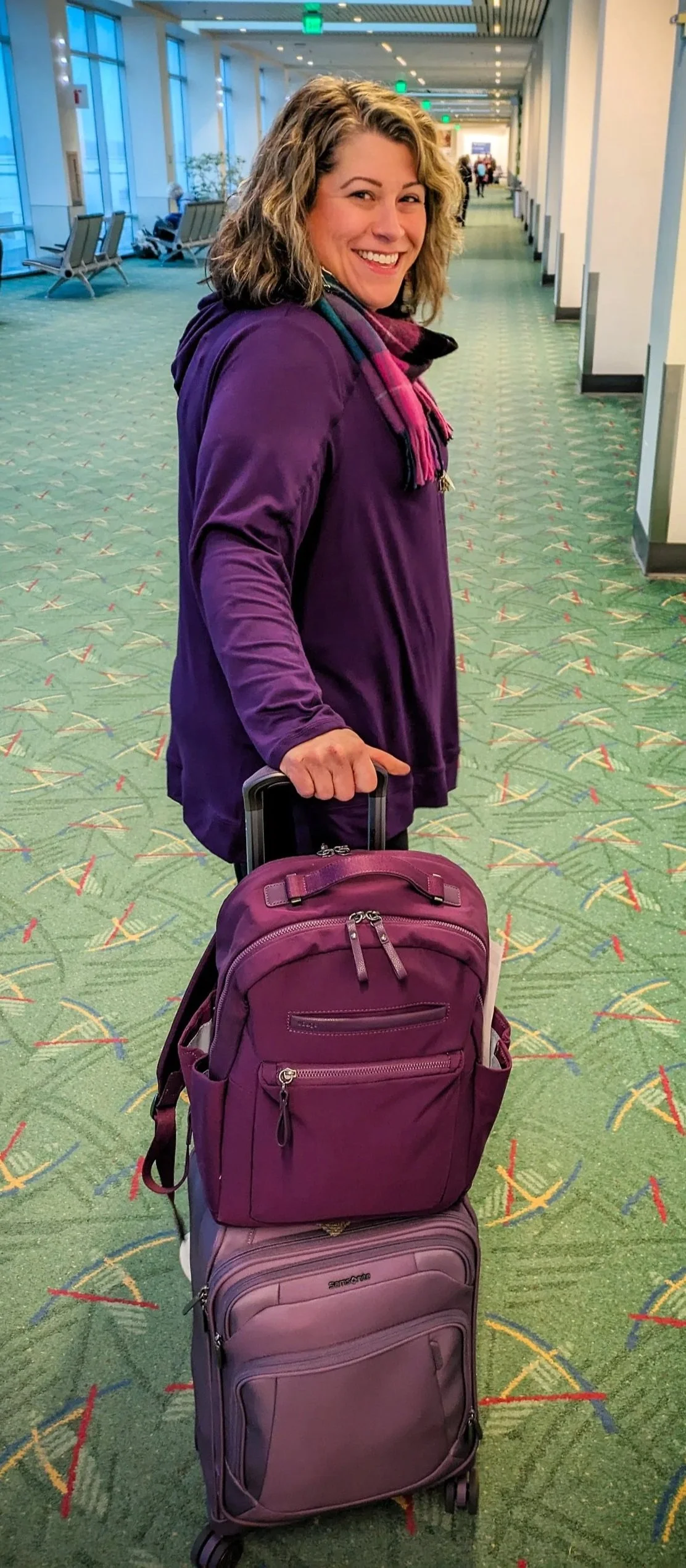 A woman with curly blonde hair, wearing a purple jacket and a multicolored scarf, is smiling and standing in an airport terminal, holding a purple rolling suitcase with a smaller purple backpack on top.
