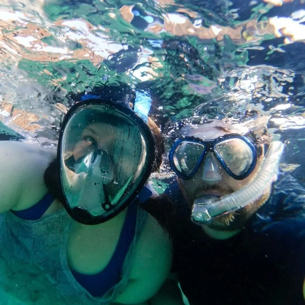 Two people snorkeling underwater, both wearing snorkeling masks and snorkels, with water above and below them.