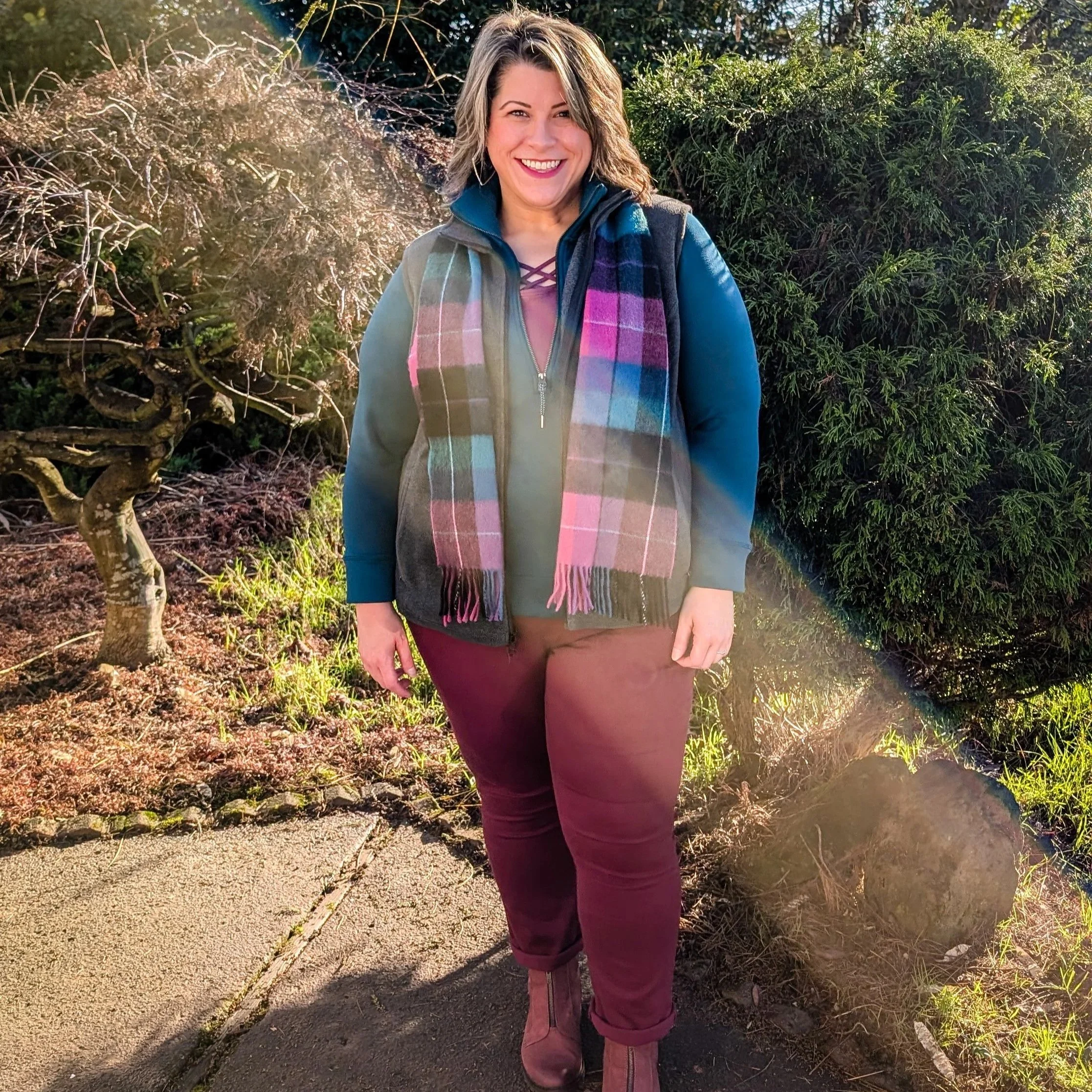 A woman smiling outdoors with trees and bushes in the background, wearing a blue jacket, colorful plaid scarf, maroon pants, and mauve boots.