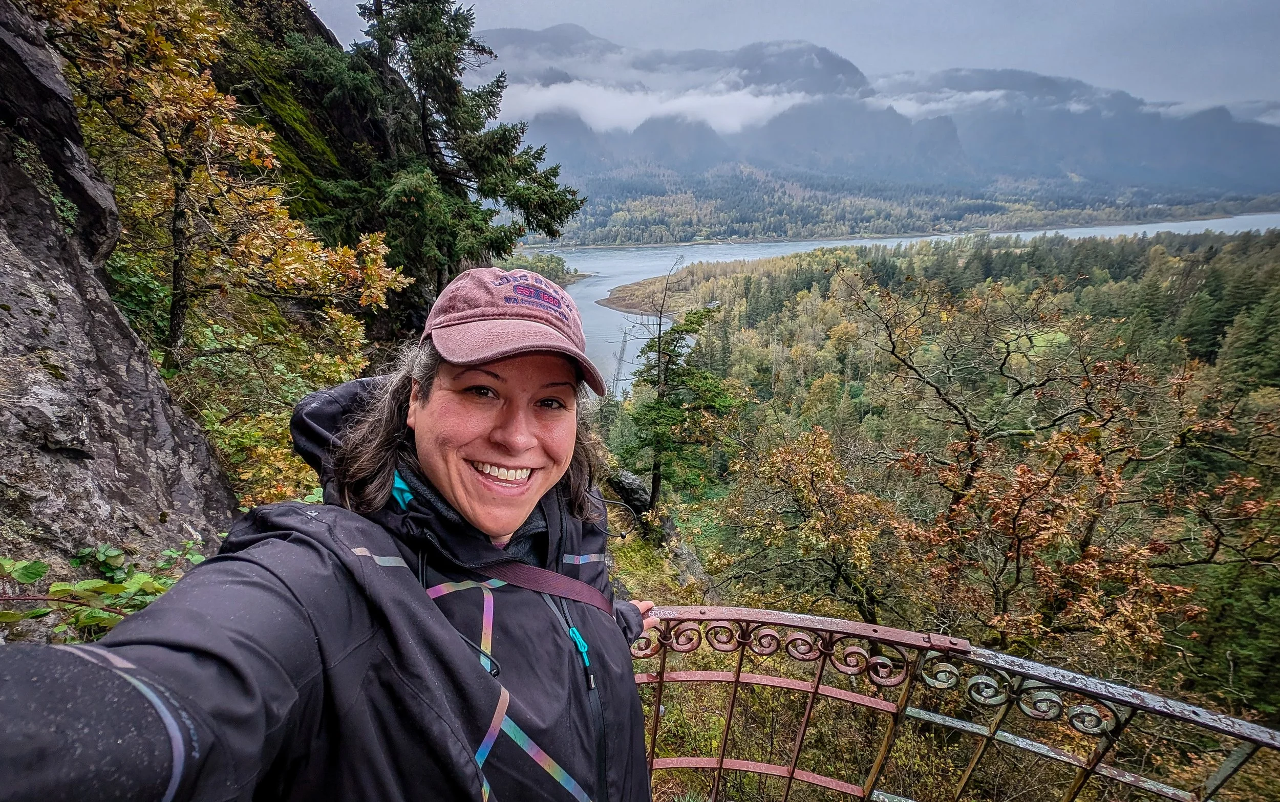 Woman in black jacket taking a selfie on a mountain trail with a view of a river, trees, and mountain range in the background, during cloudy weather.