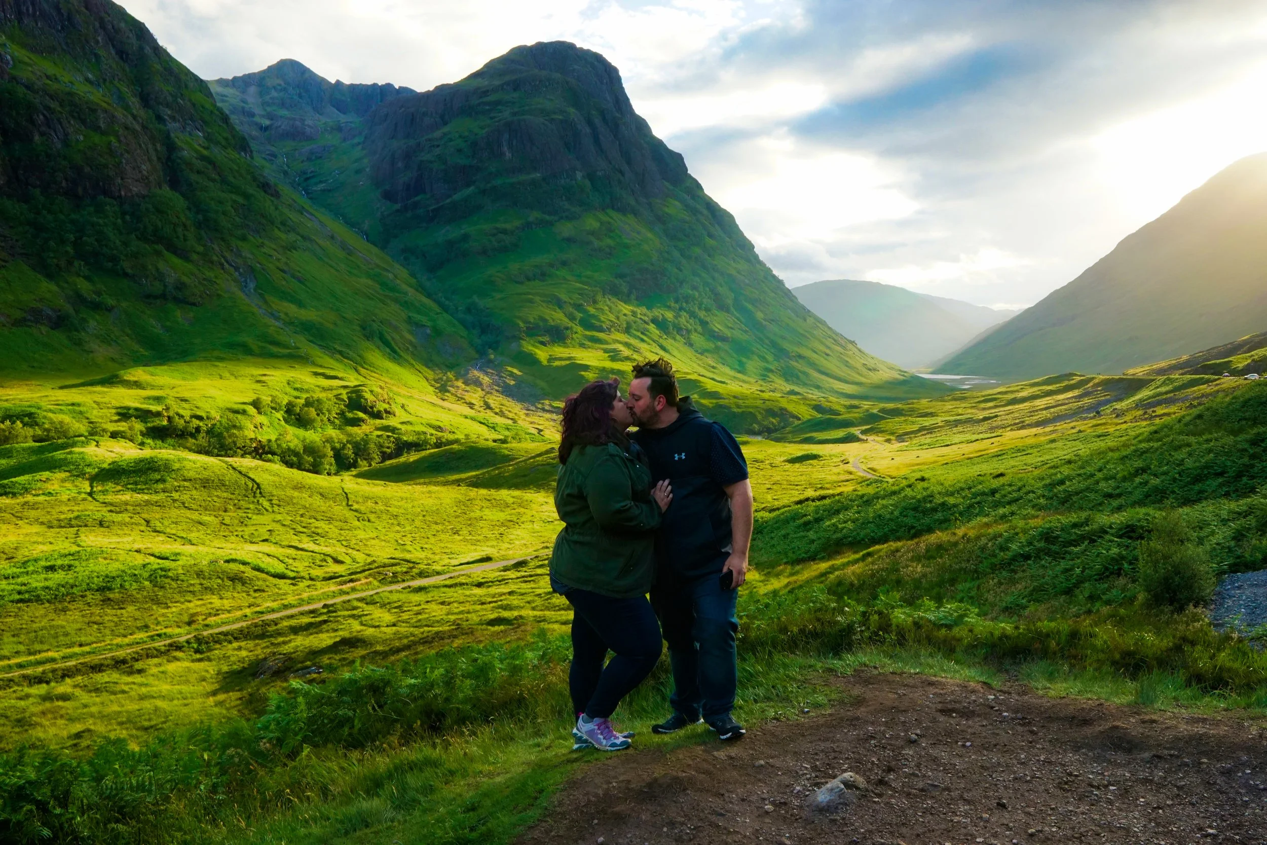 Couple kissing in a lush green valley surrounded by mountains at sunset.