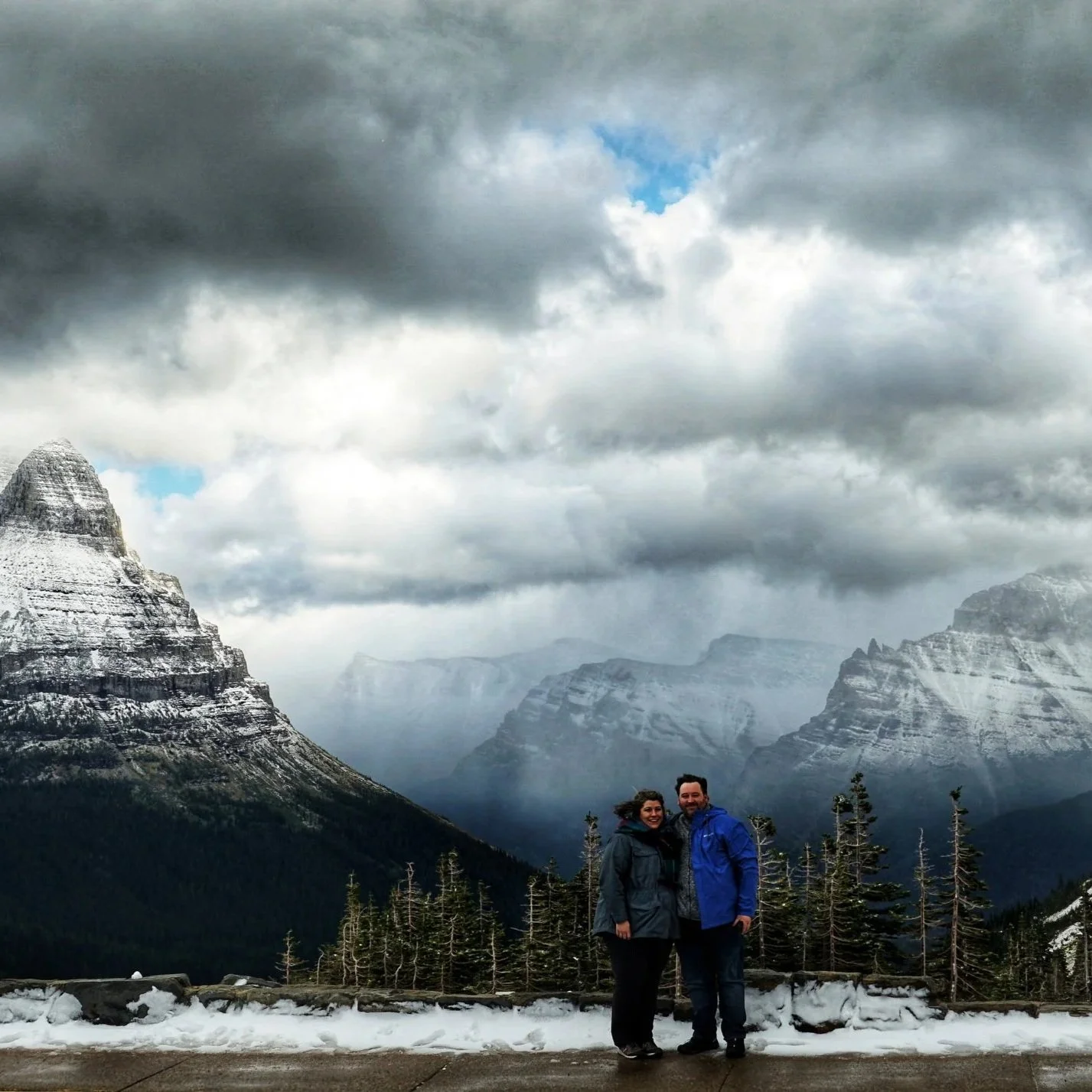 A couple standing on a snowy path with mountains, pine trees, and dark clouds in the background.