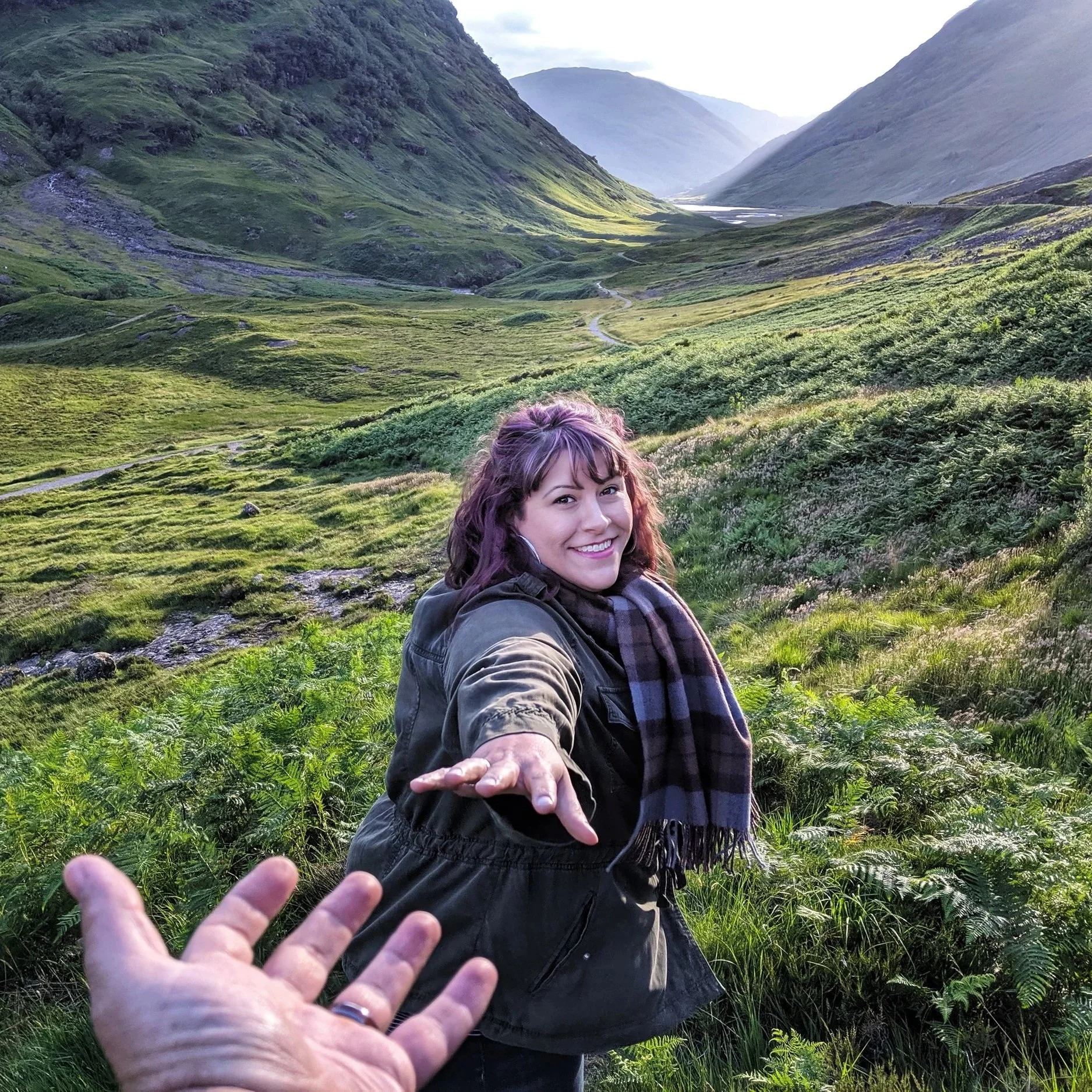 A woman smiling and reaching out with her hand in a lush green valley with mountains in the background.