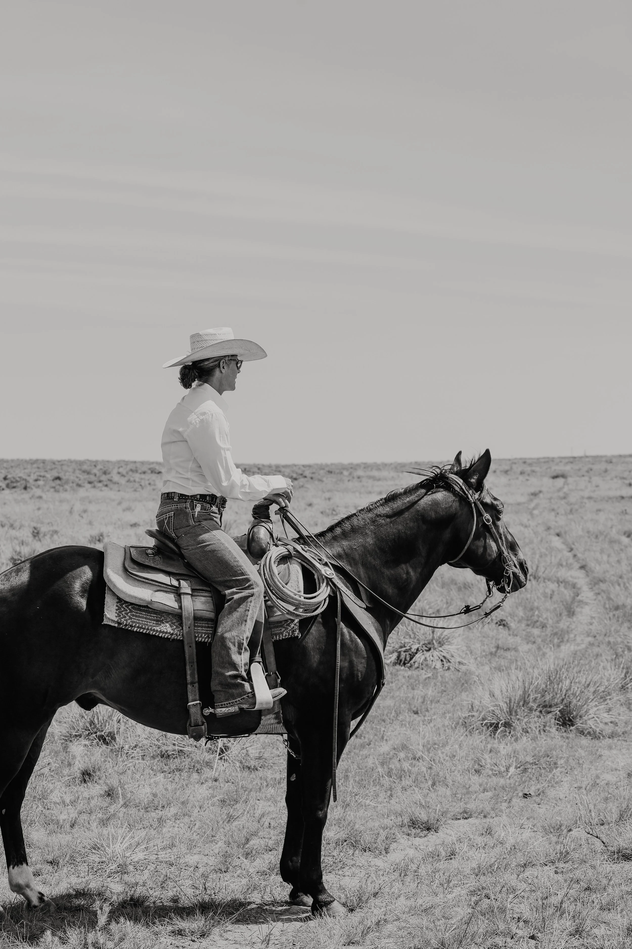 A woman in a white shirt, jeans, and a wide-brimmed hat riding a horse across an open field with tall grass under a cloudy sky.