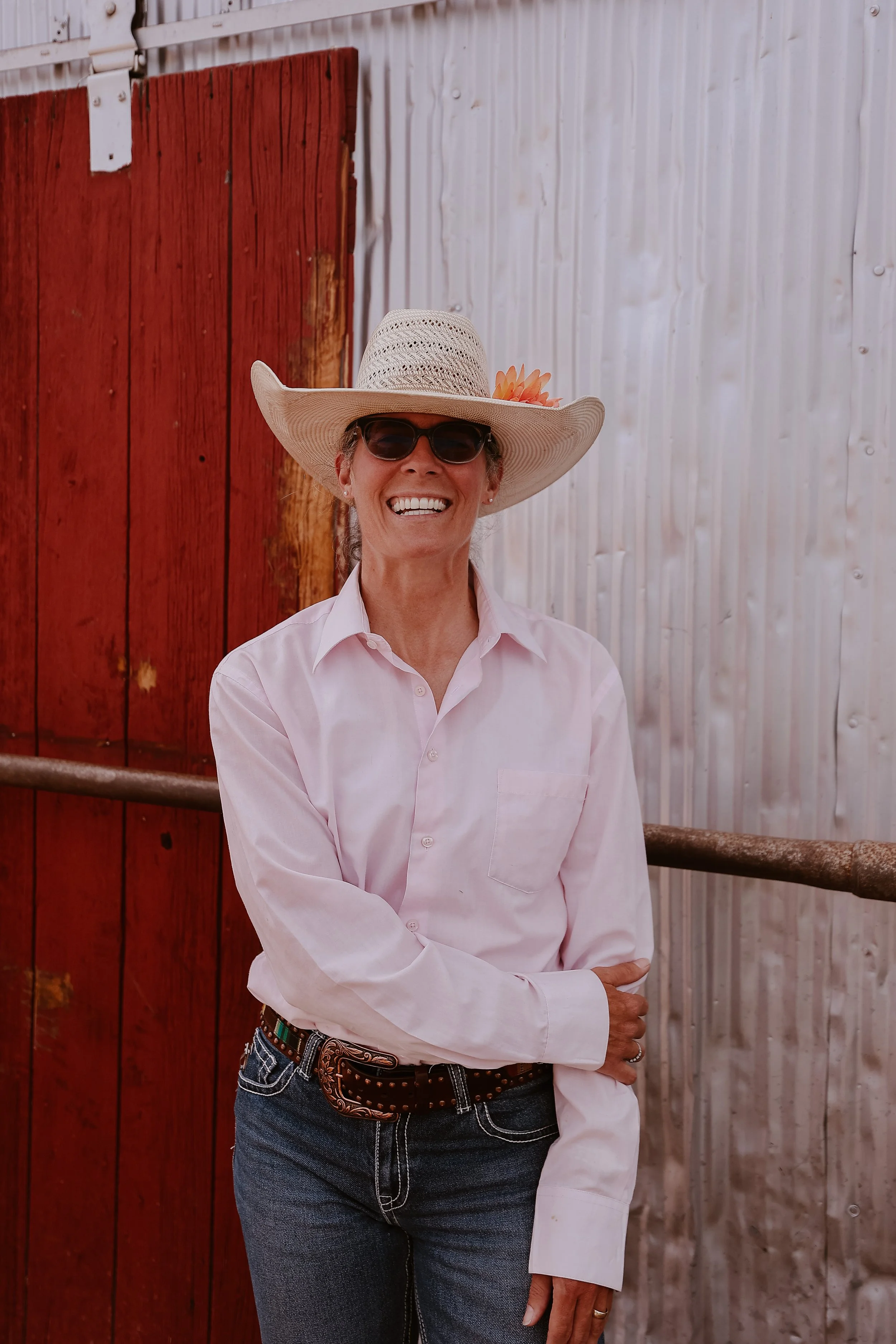 A woman wearing a white shirt, sunglasses, and a large straw hat with a pink flower, standing against a red and gray wooden wall, crossing her arms and smiling.