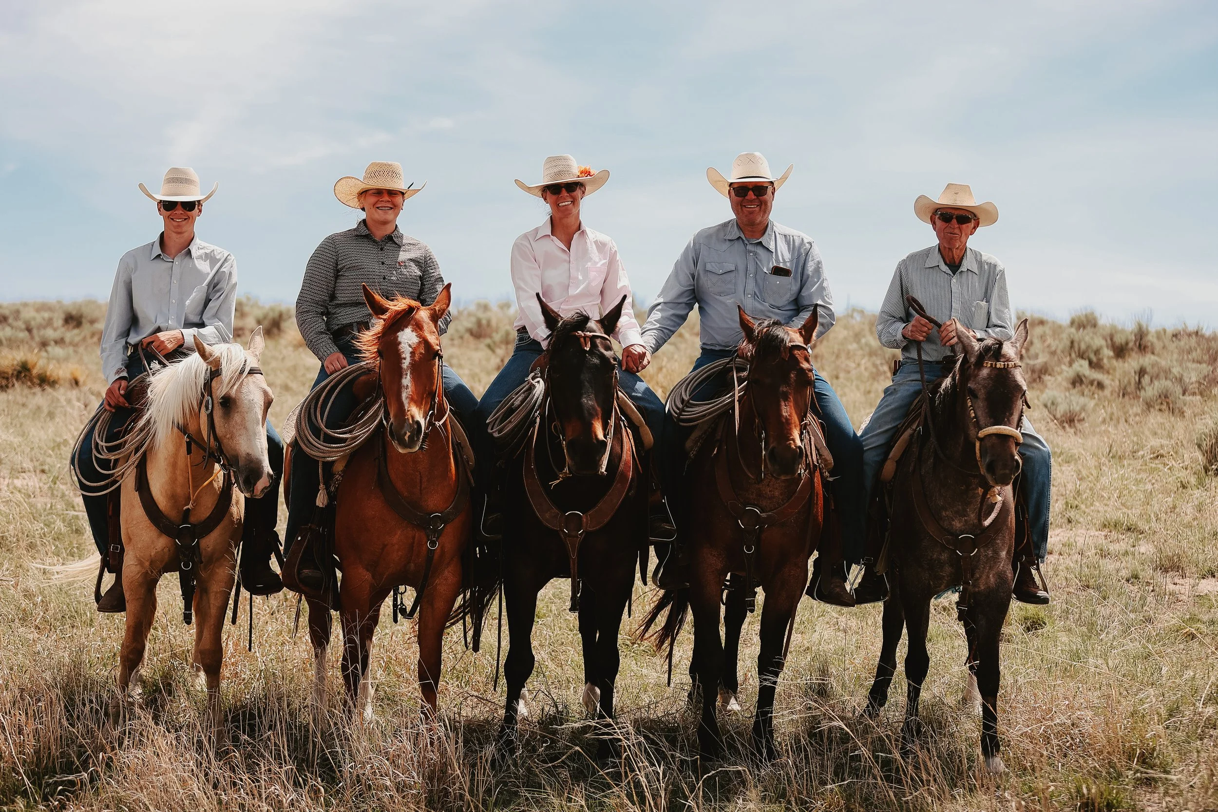 Group of five people riding horses in an open field, wearing cowboy hats and casual shirts, smiling under a partly cloudy sky.