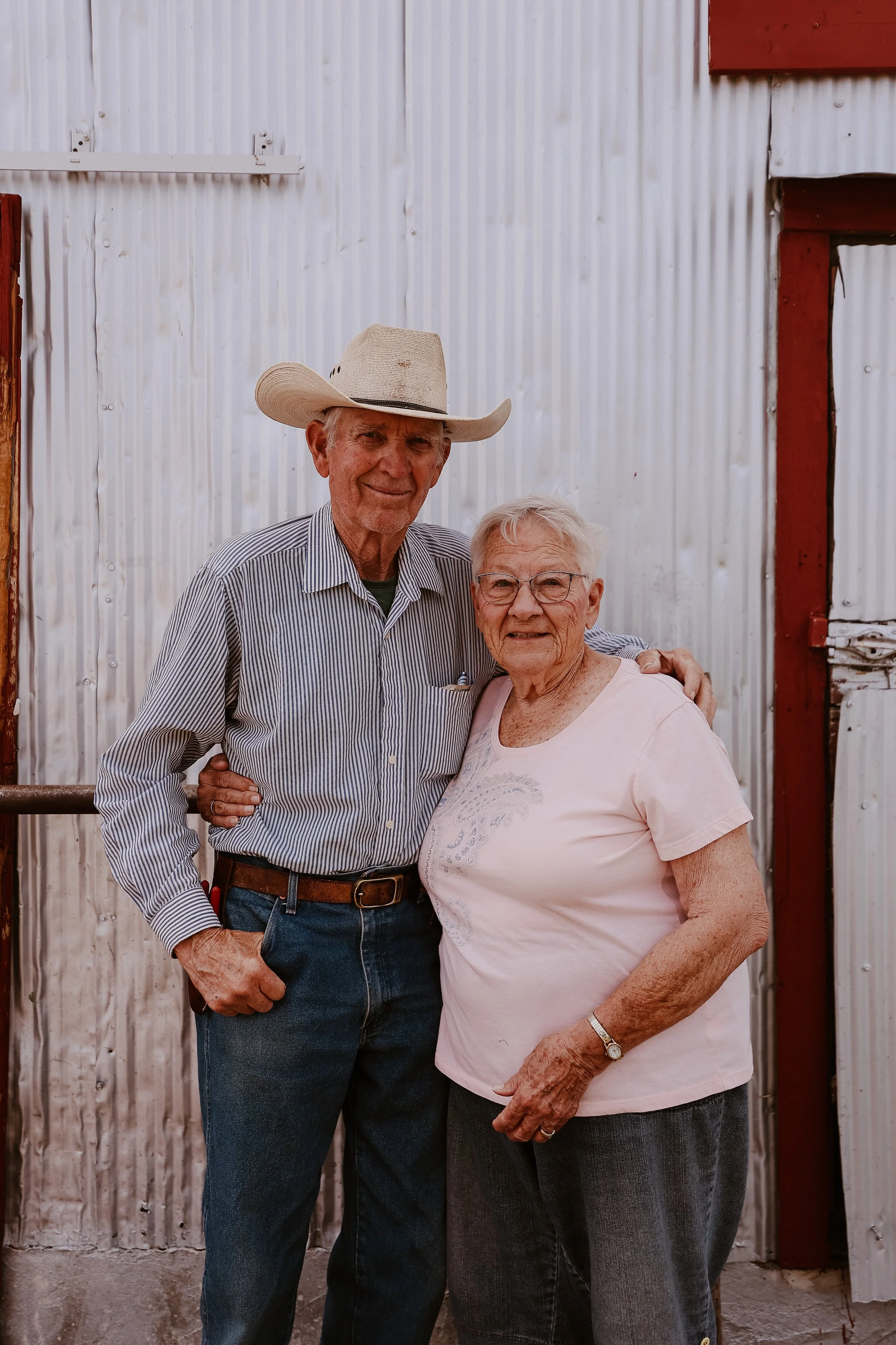 An elderly man and woman standing outdoors in front of a corrugated metal wall, smiling and embracing.