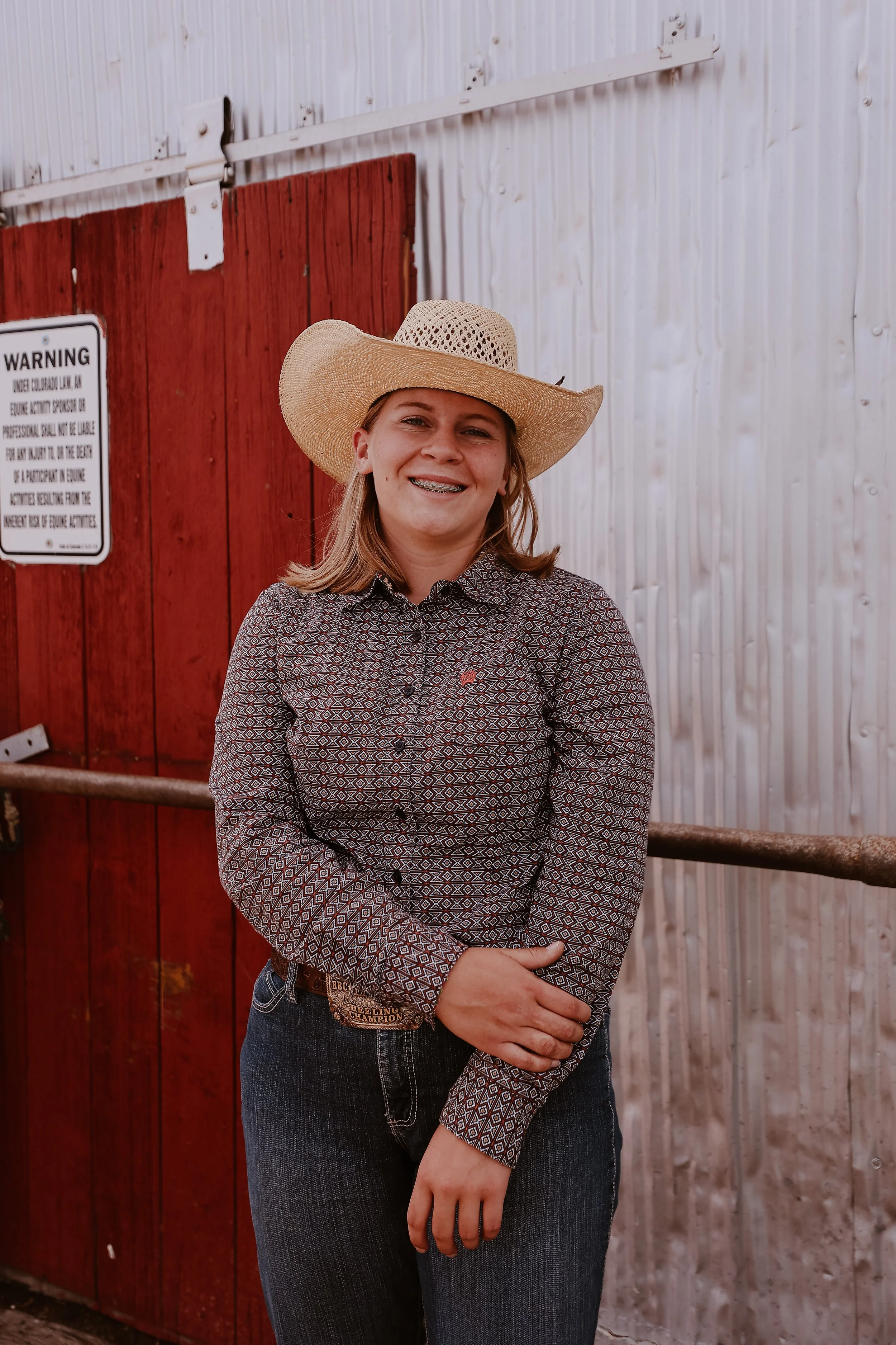 A young woman with blonde hair wearing a wide-brimmed cowboy hat, a patterned long-sleeve shirt, and blue jeans, standing in front of a wooden fence with a red barn door and a warning sign.