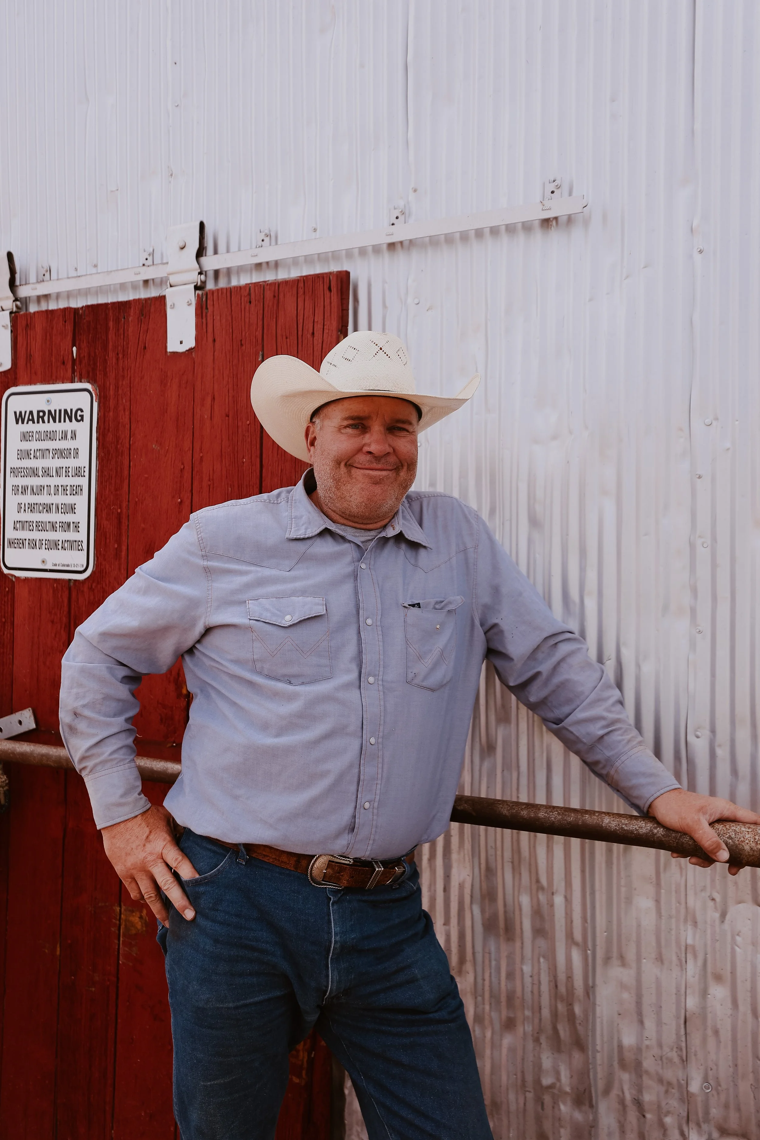 A man wearing a white cowboy hat, light gray shirt, and blue jeans standing outdoors with his right hand resting on a brown metal pipe in front of a wooden and metal background. There is a sign on the red wooden fence that provides a warning about liability in equine activities.