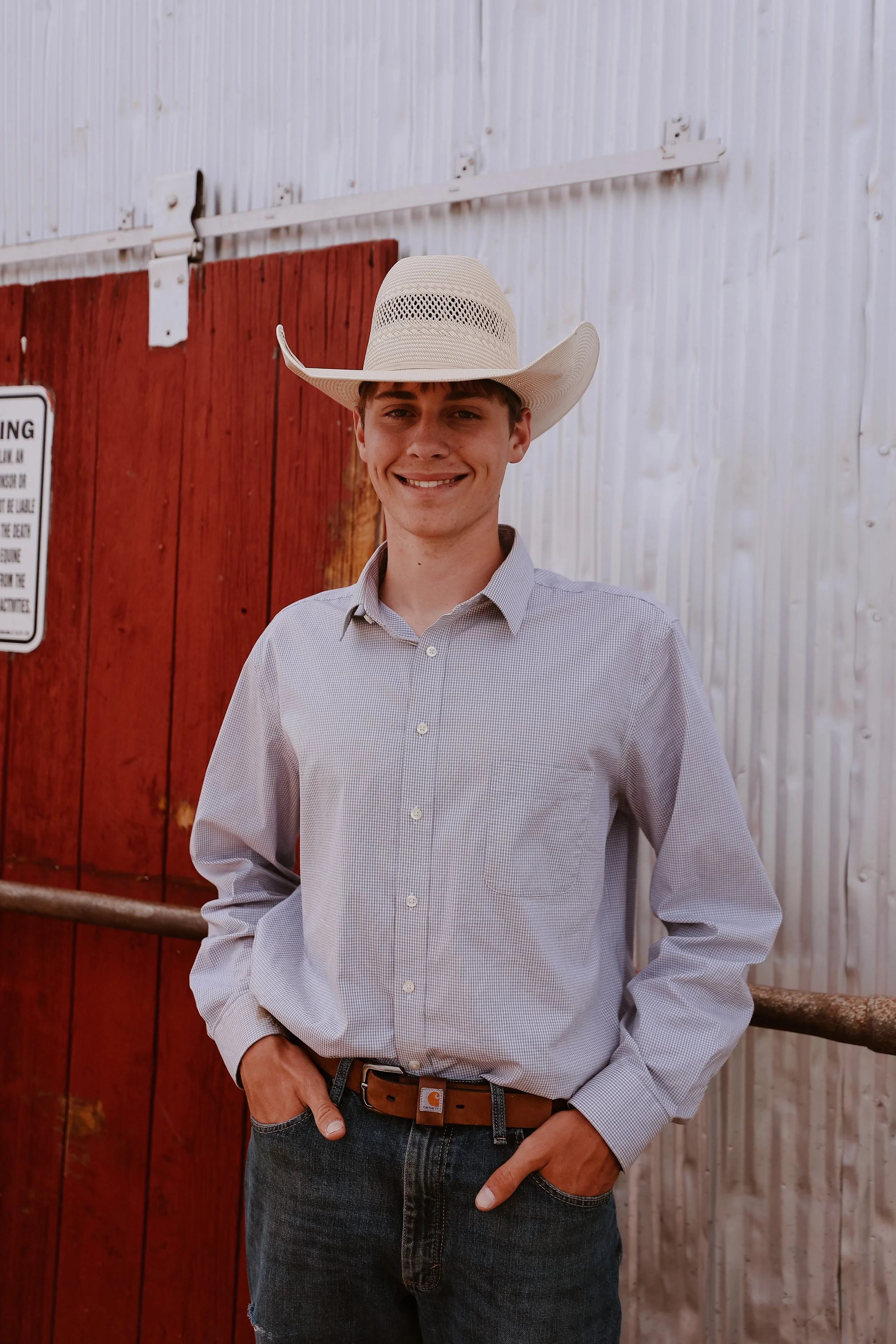 A young man wearing a cowboy hat, a light gray checkered shirt, and dark jeans, standing outdoors in front of red and white barn doors, smiling at the camera.