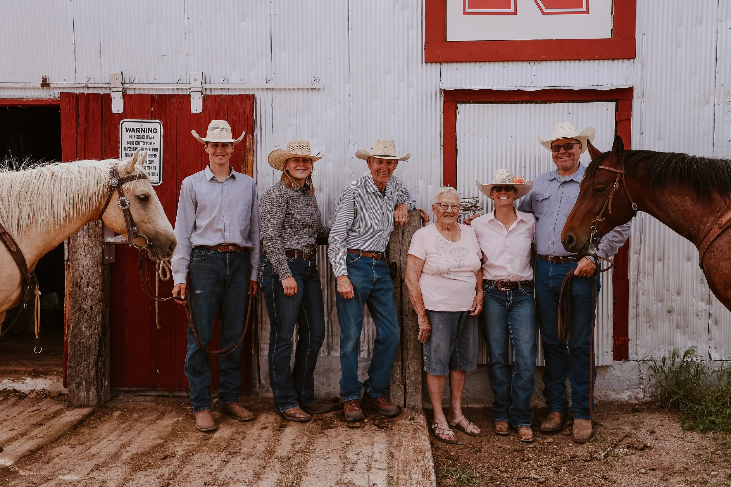 Group of seven people, including men and women of different ages, standing outside a barn with white corrugated metal walls and red trim. Two of the men and one woman are holding horses, and each person is wearing a cowboy hat. They are smiling and posing for the photo.