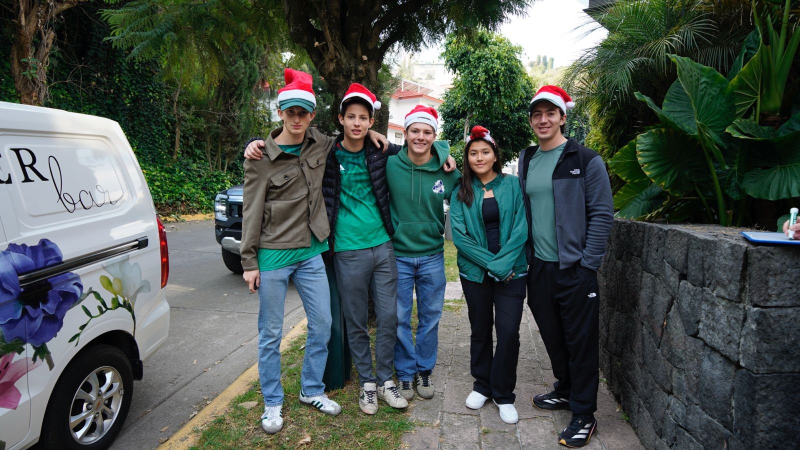 Five young people standing together outdoors, dressed in casual clothing and holiday Santa hats, smiling and posing for the photo.