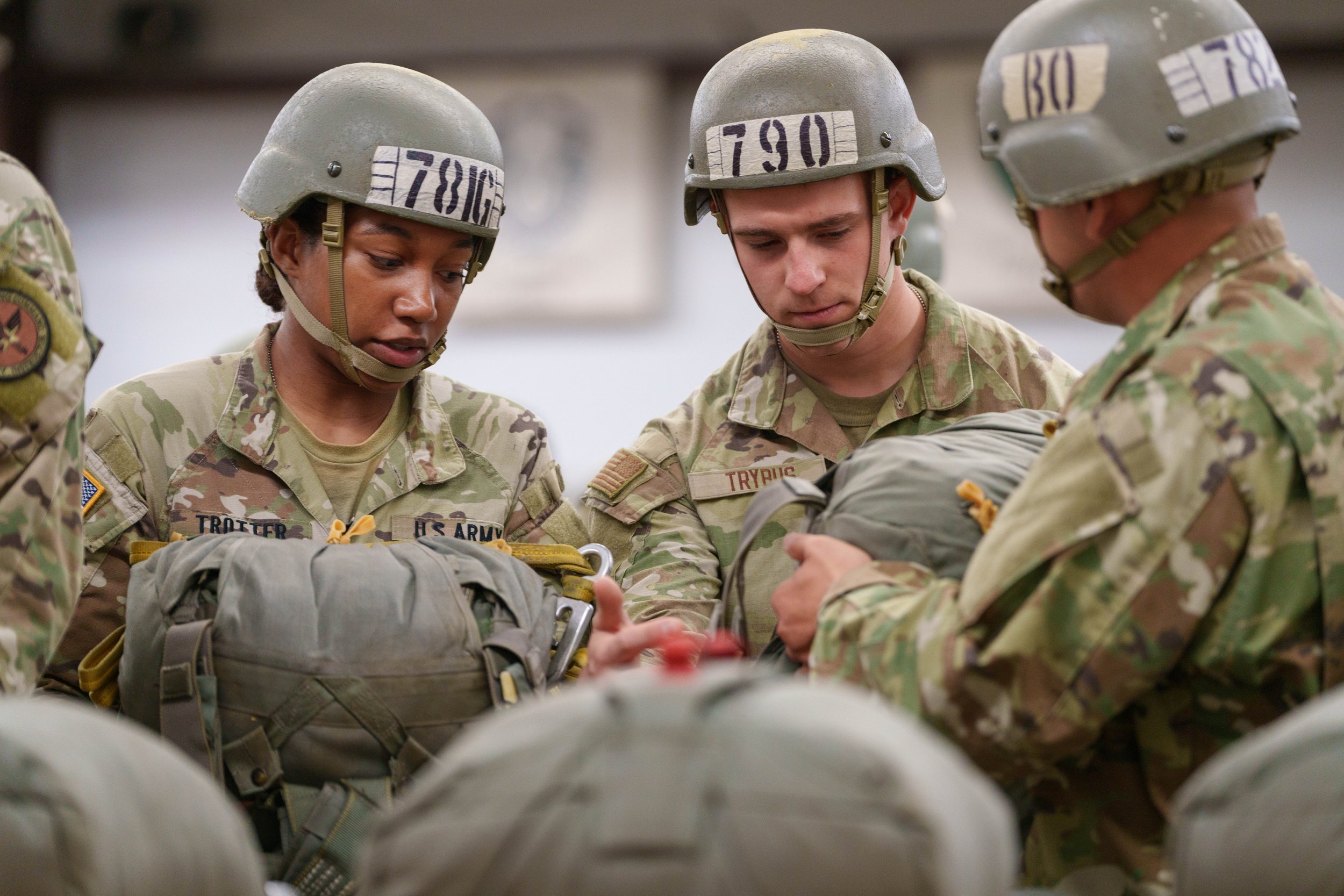 Three soldiers in camouflage uniforms and helmets looking at their gear, with one soldier showing something to the others.