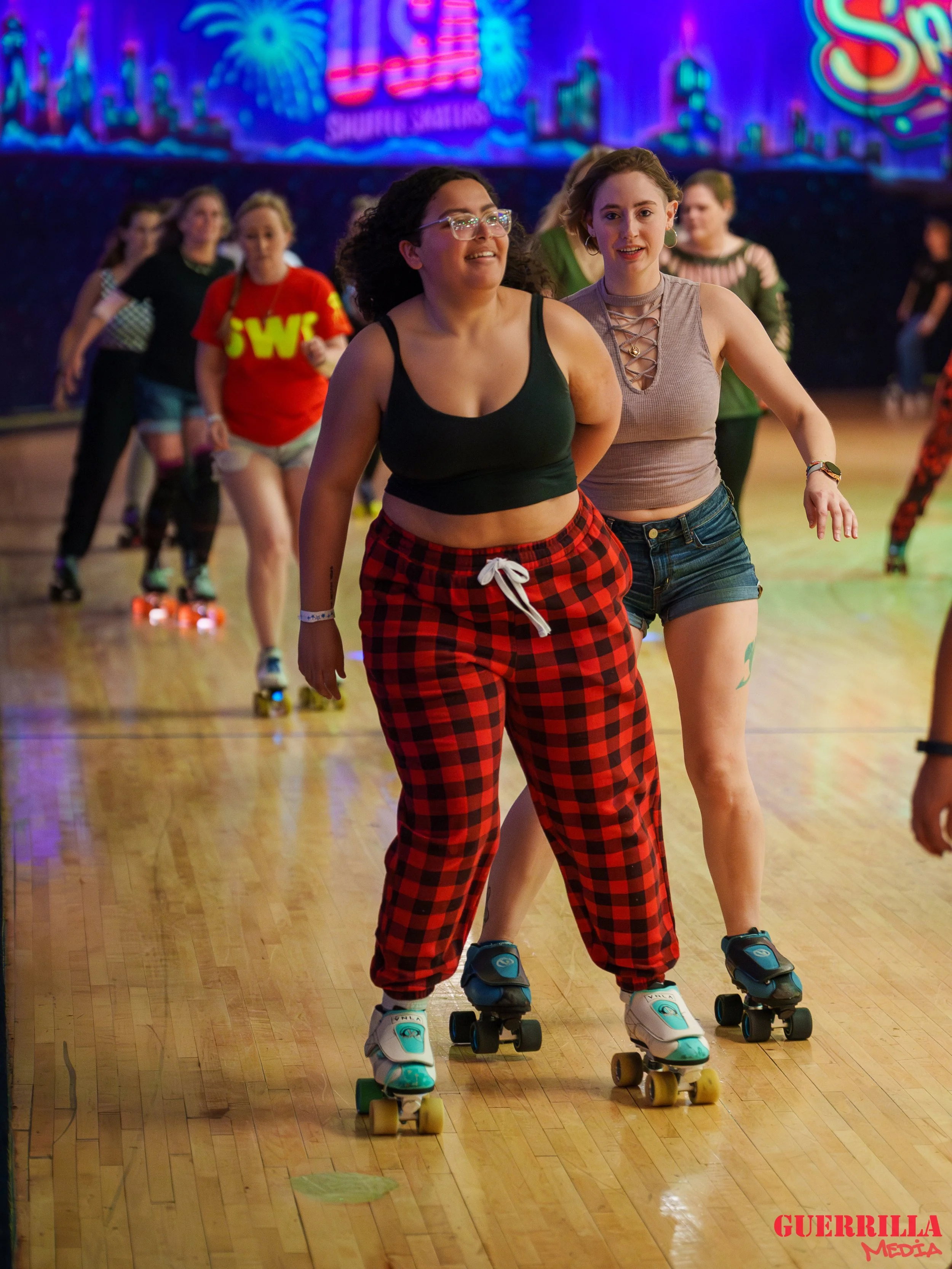 Two women roller skating at an indoor roller rink, with colorful neon decorations in the background and other skaters in the distance.