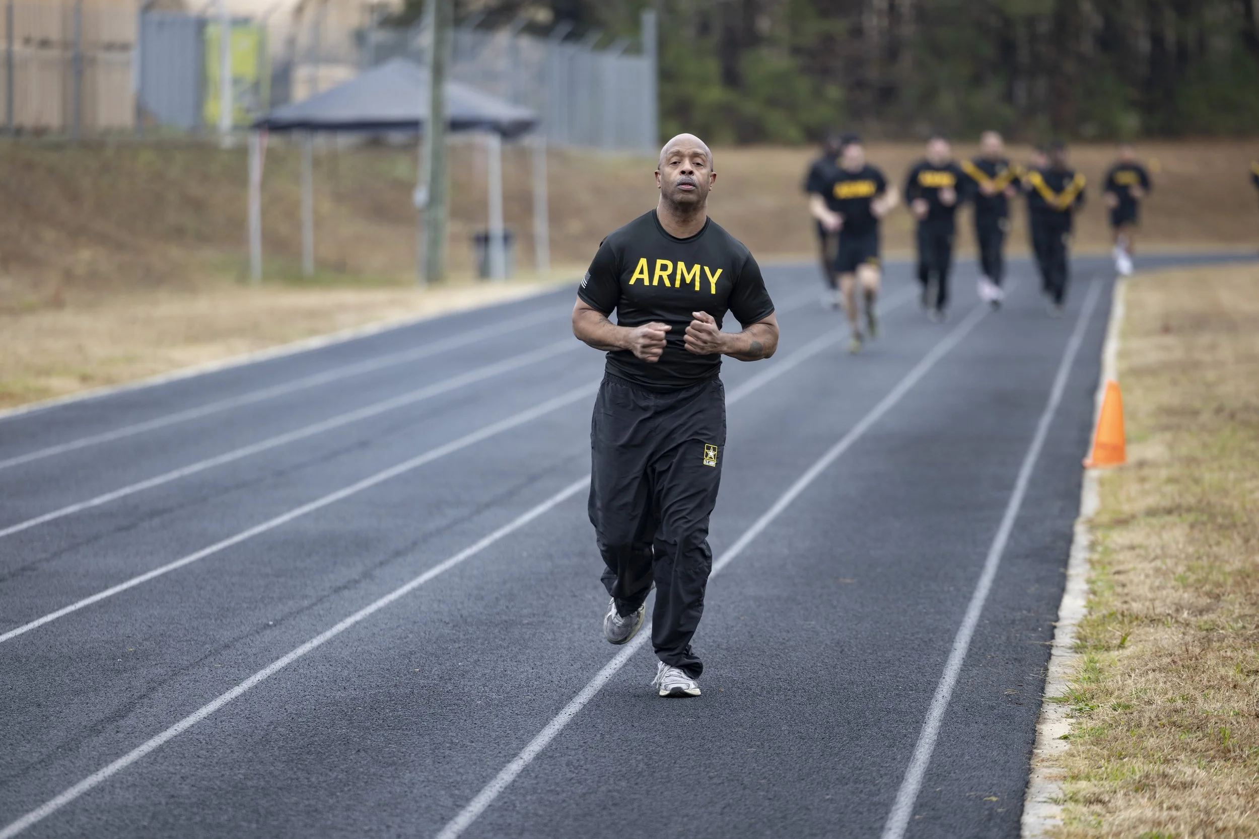 A man in military training attire running on a track with other runners in the background. He is wearing a black shirt with 'ARMY' written on it and black pants.