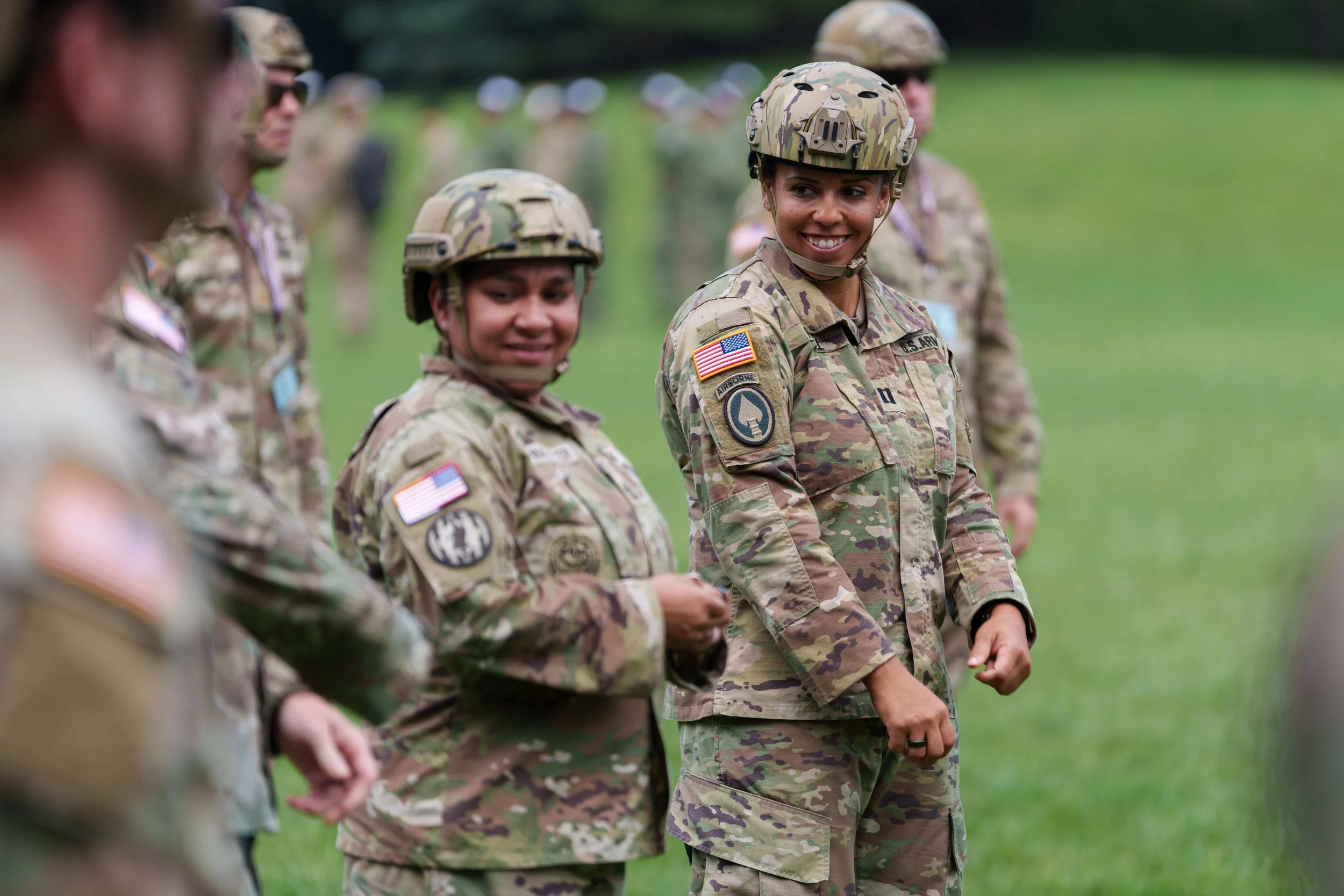 Group of female soldiers in camouflage uniforms and helmets standing outdoors on a grassy field, smiling and engaging with each other.