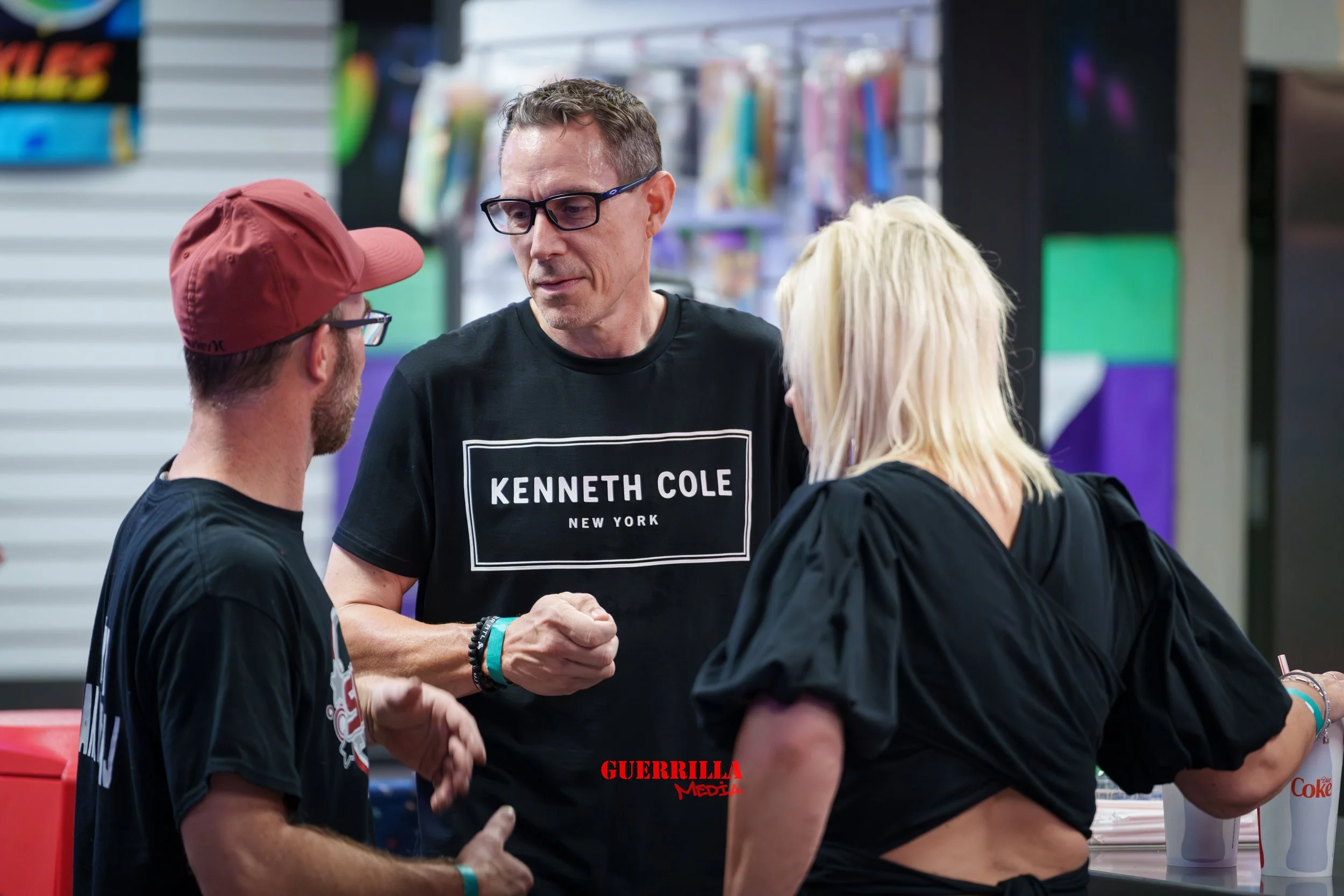 Three people engaged in conversation at a counter inside a store or arcade, with colorful prizes or merchandise in the background.