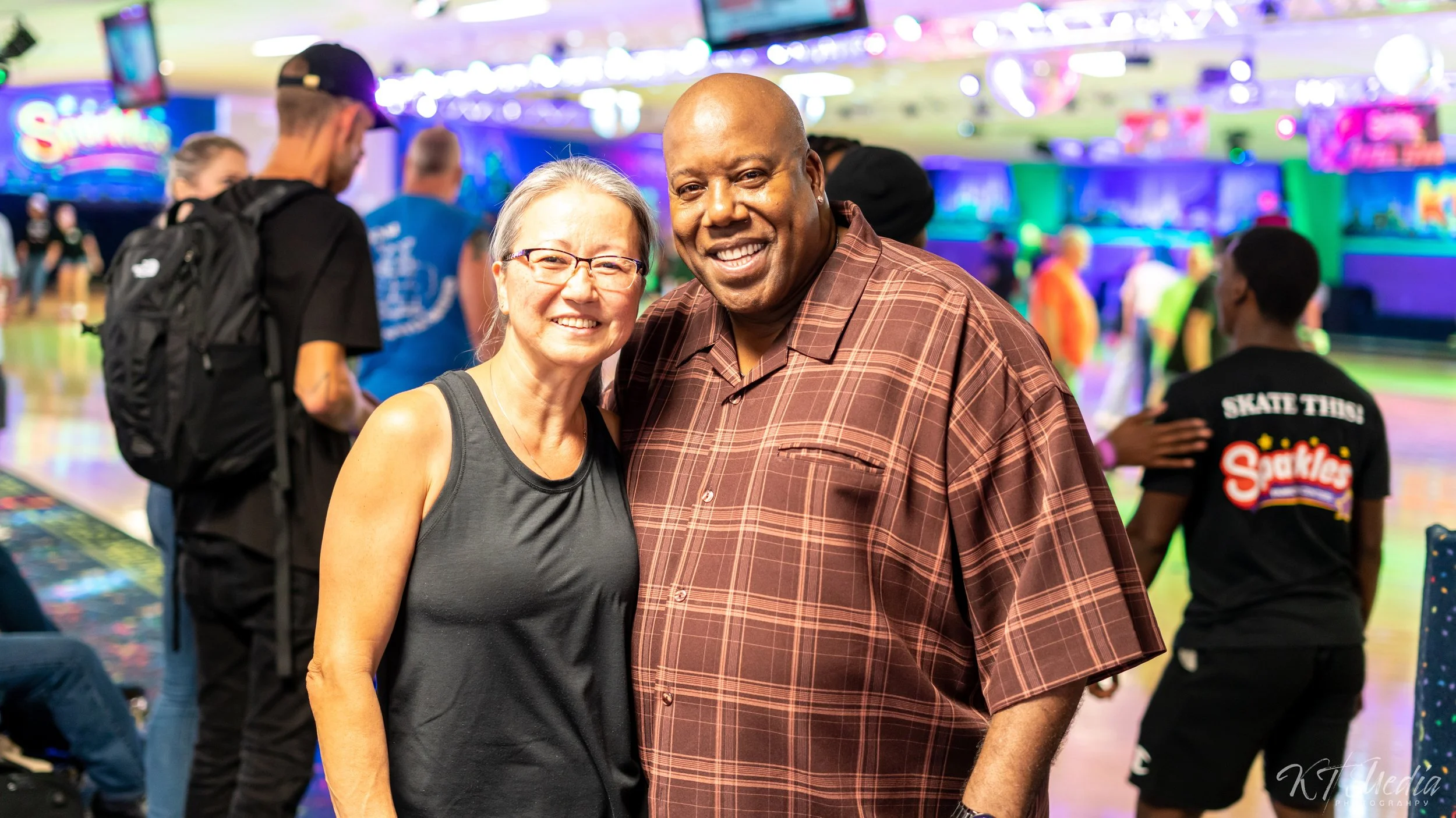 A smiling woman and man pose for a photo at a colorful indoor skating rink. The background shows other people, bright lights, and skating area.