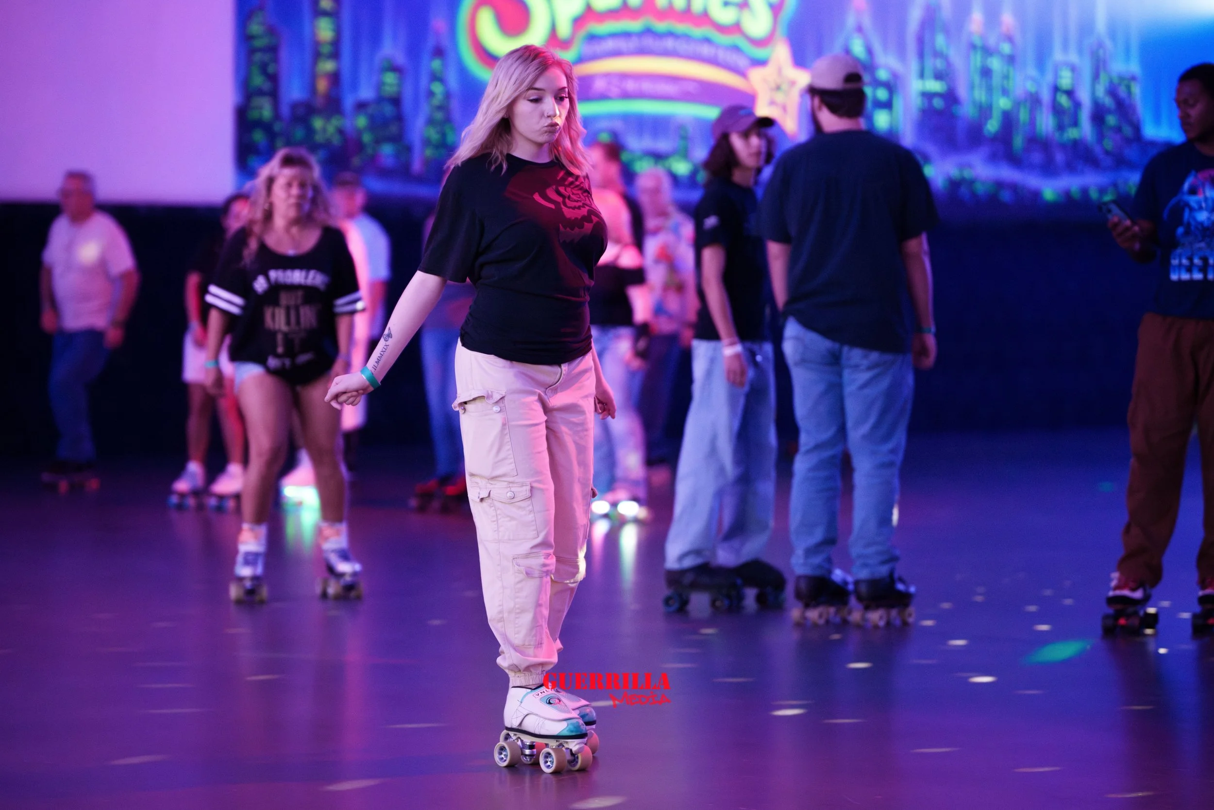 A young woman with blonde hair roller skating in an indoor roller skating rink with other skaters. The background has colorful neon signs and cityscape graphics.