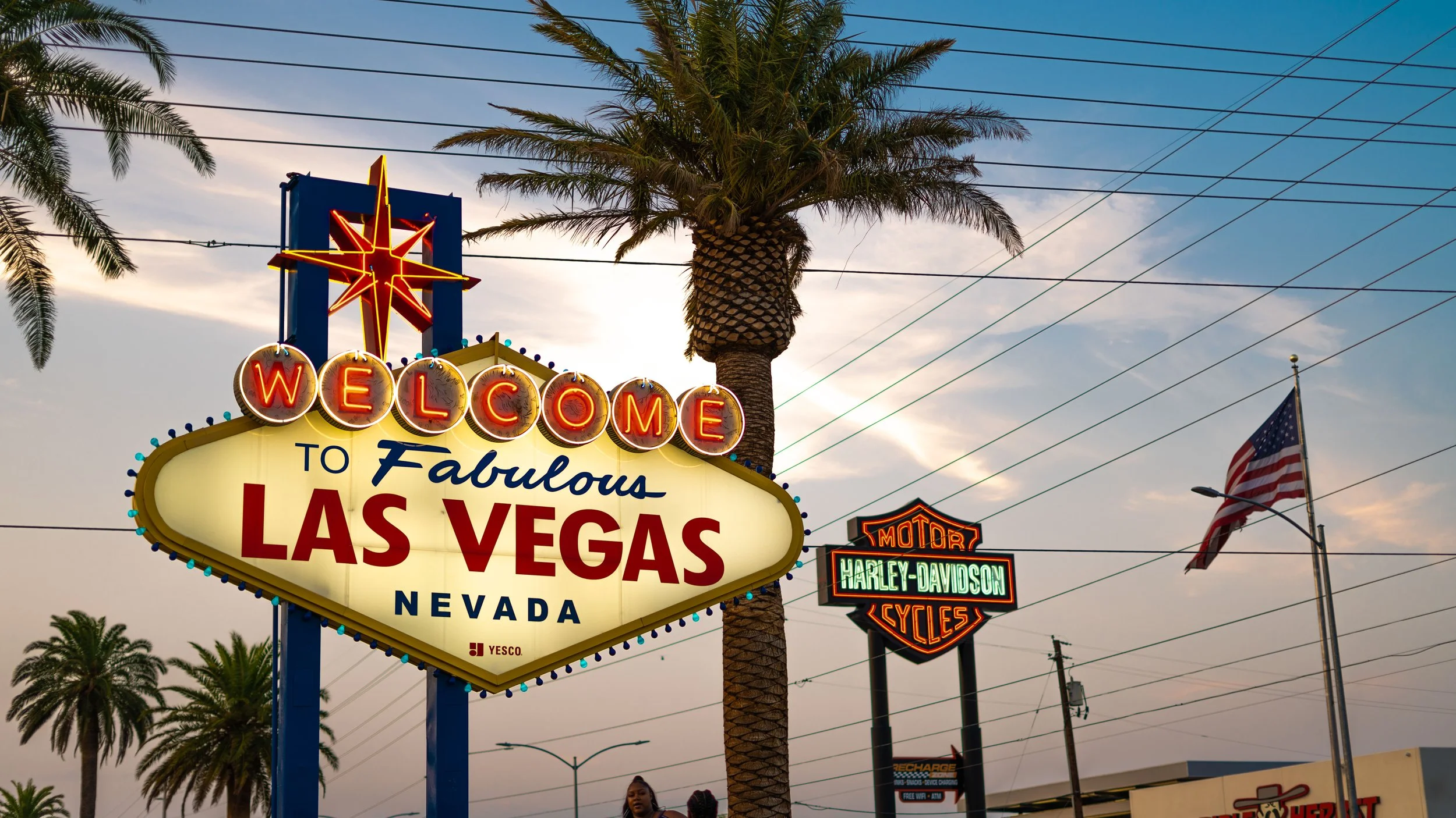 The famous 'Welcome to Fabulous Las Vegas Nevada' sign at dusk, with neon lights illuminated, a palm tree, an American flag, and a Harley-Davidson sign in the background.