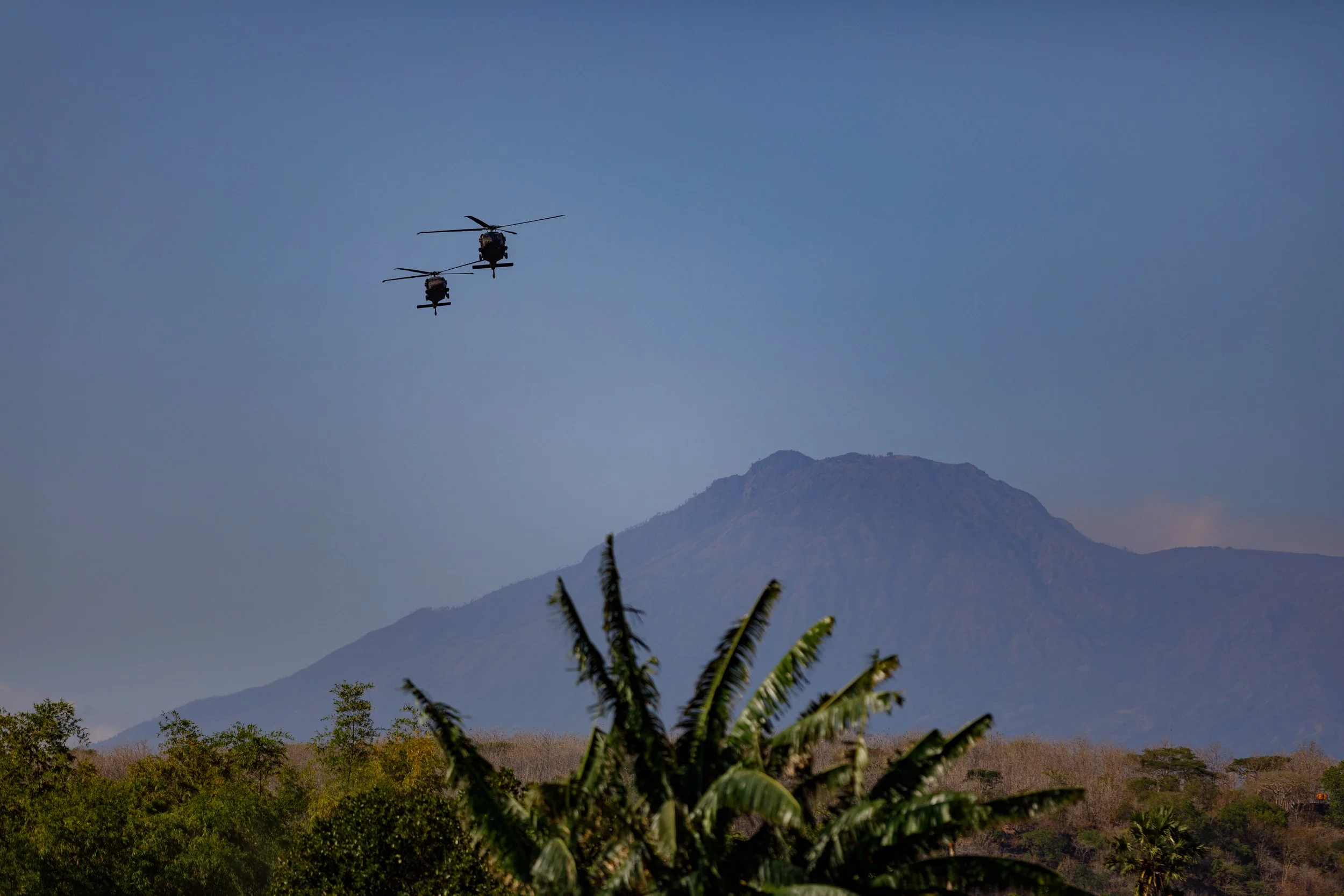 Two helicopters flying above a mountainous landscape with trees and a volcano in the background.