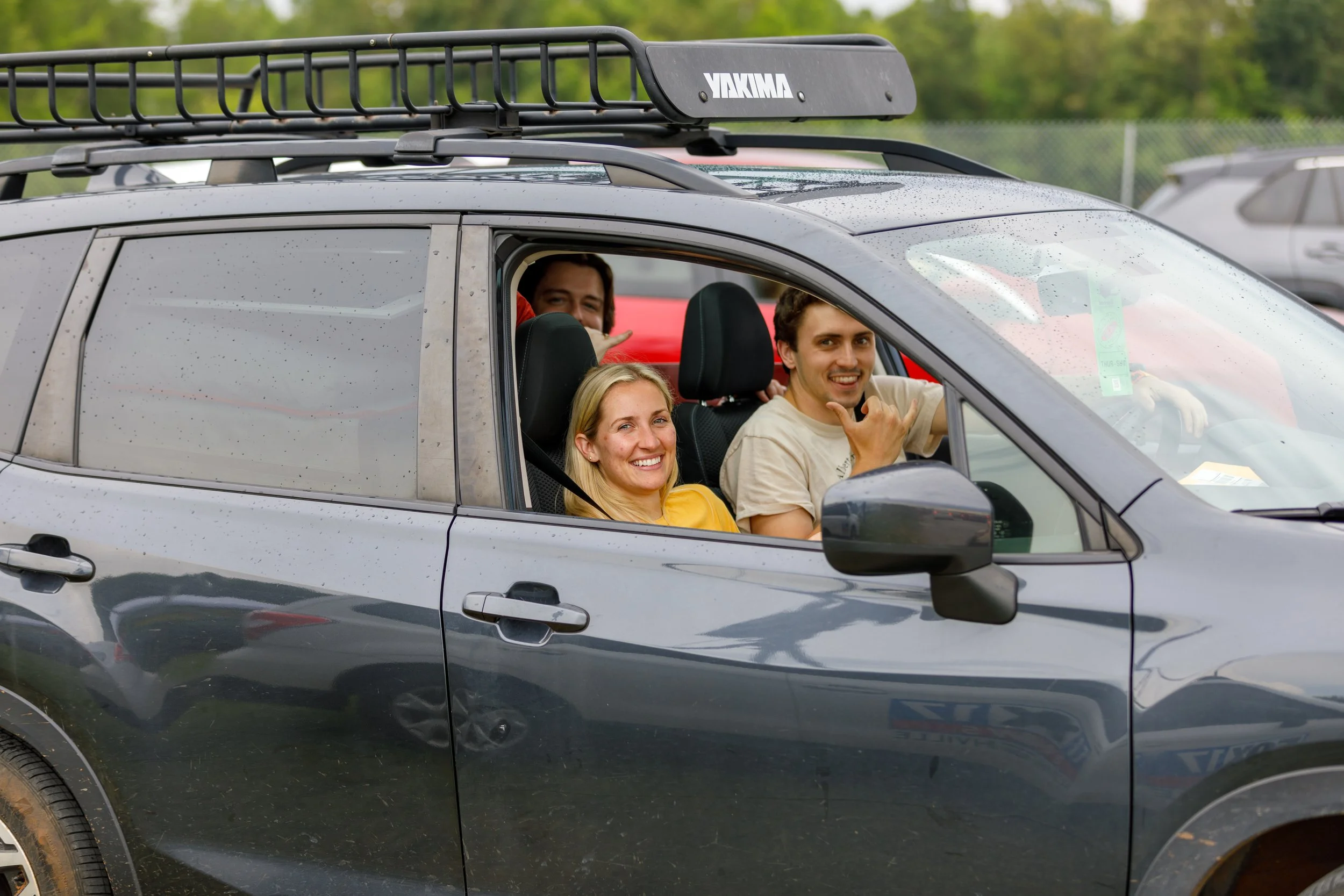 Four people sitting inside a gray SUV with an outdoor parking lot in the background. The front passenger seat is occupied by a smiling blonde woman, and beside her the young man in the driver's seat is making a hand gesture. Two more people are in th