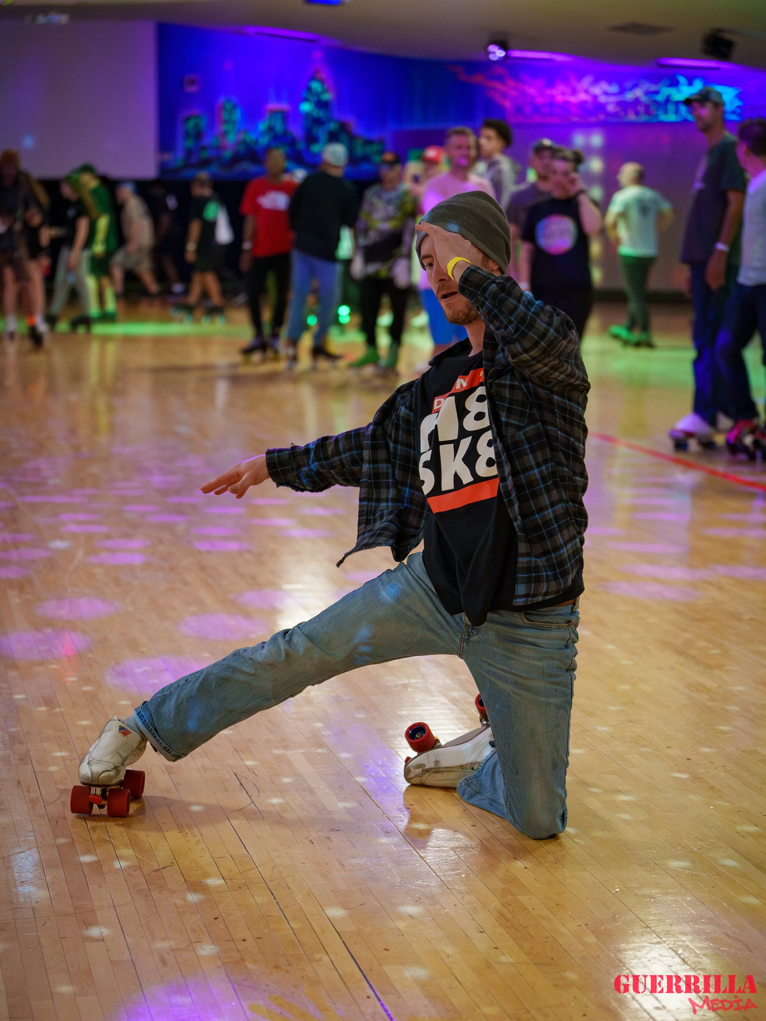 A man roller skating indoors on a wooden floor, one knee on the ground and the other leg extended out, with a hand on his head in a dramatic pose, while a large group of people watch in the background at a roller skating rink.