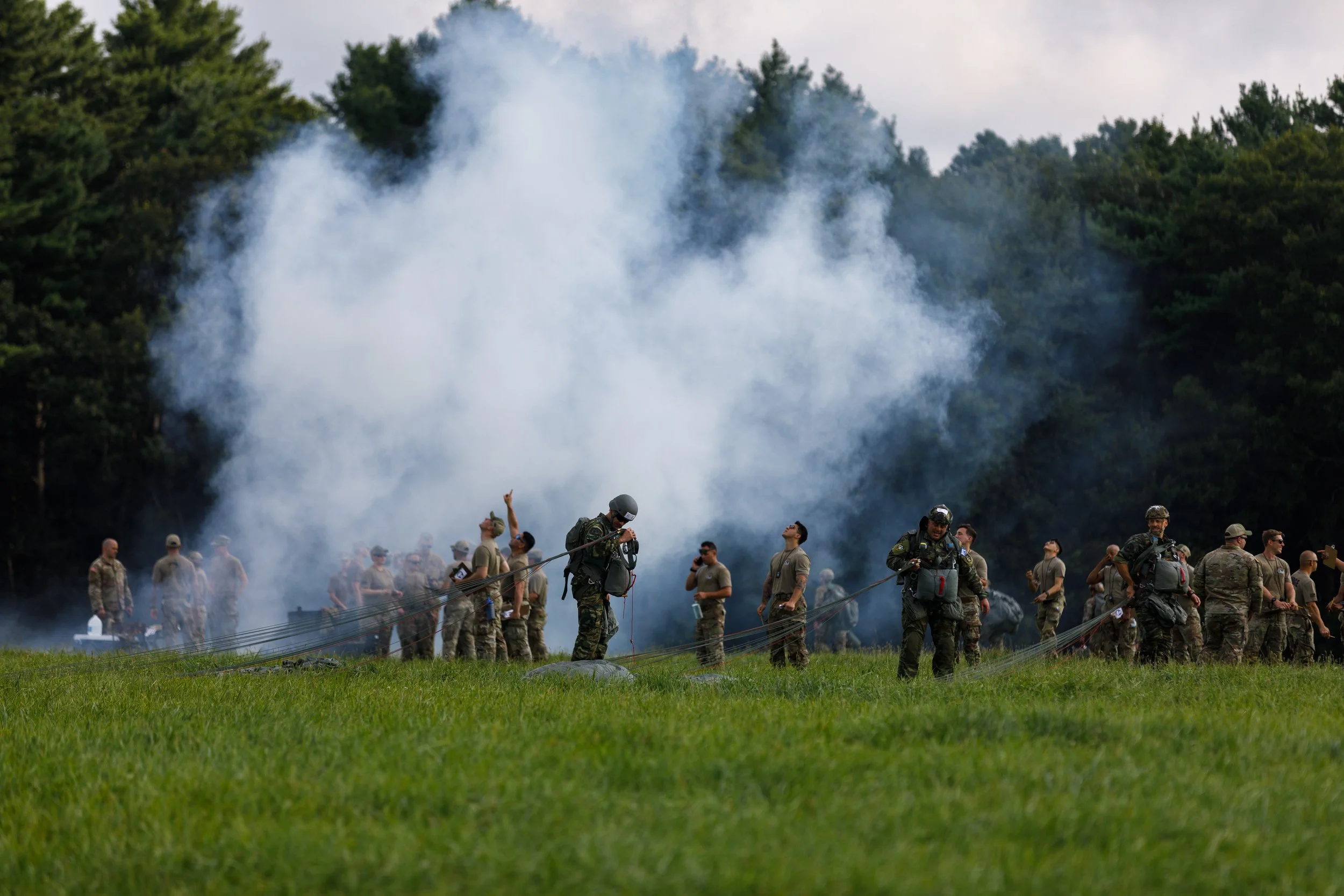 A group of soldiers standing outdoors on a grassy field with trees in the background, some holding or adjusting parachutes while smoke rises behind them.