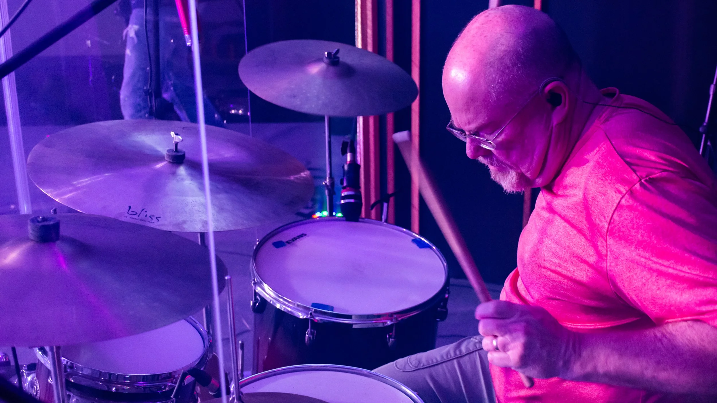 A man with glasses and a beard playing drums on stage under purple lighting, with cymbals and drumsticks visible.