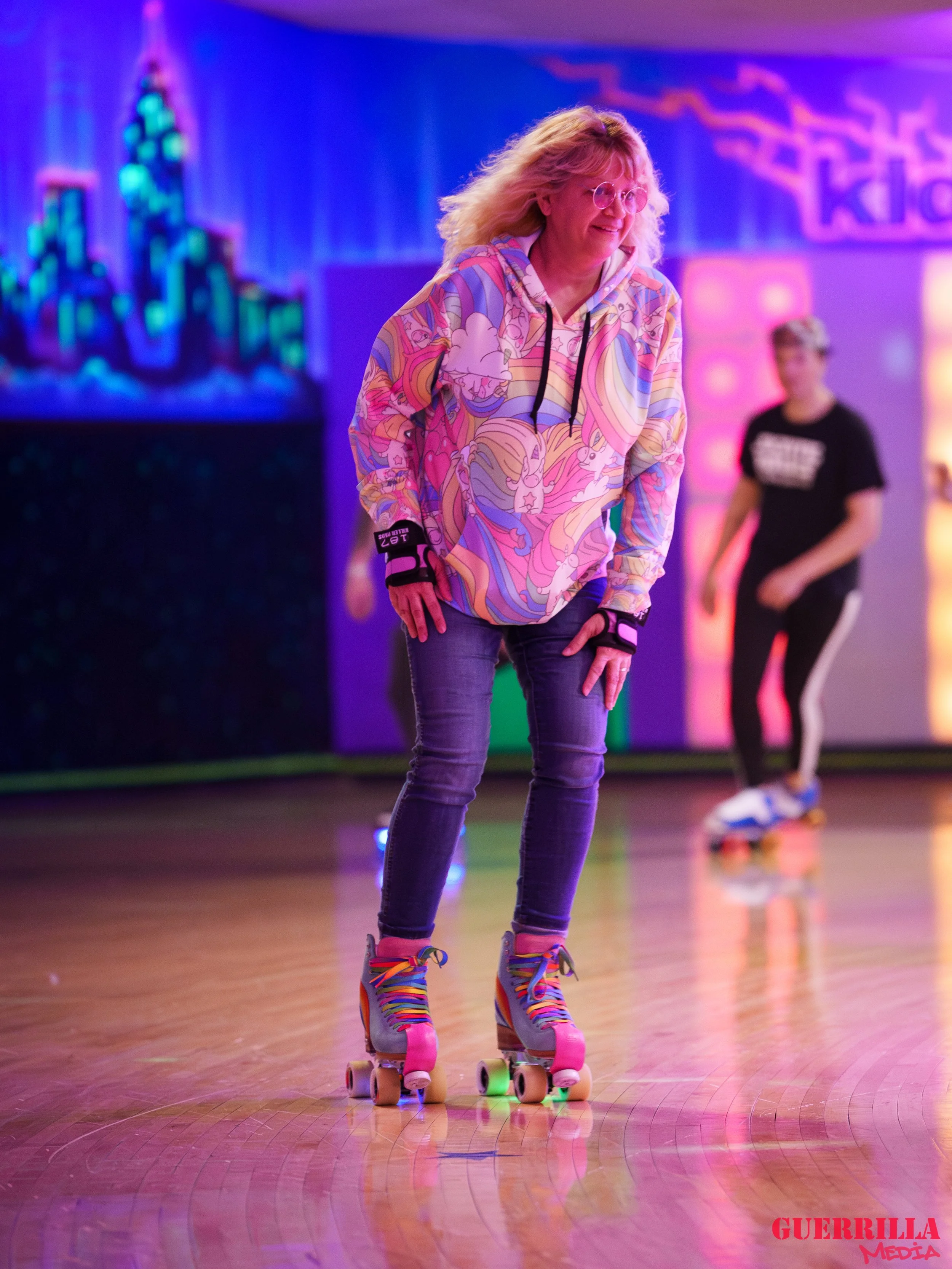 A woman wearing a colorful hoodie and roller skates gliding on a roller rink with neon lights and other skaters in the background.