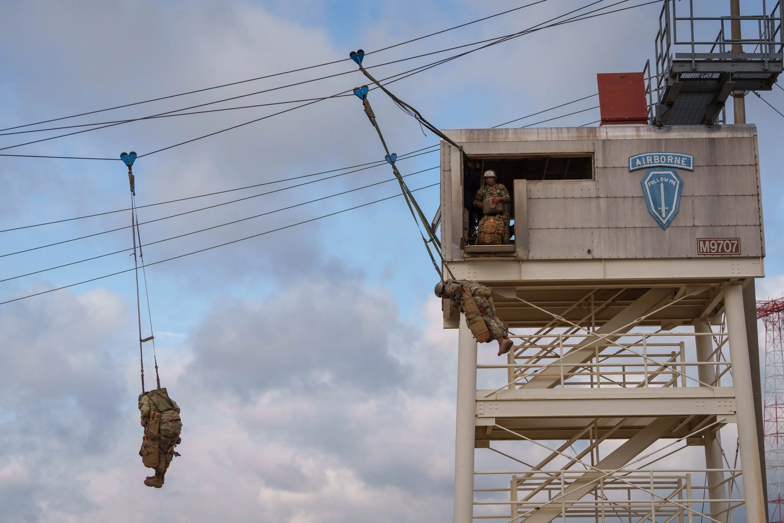 Military personnel practicing rope rescue training on a tower, with one soldier rappelling down and others watching from the observation deck.