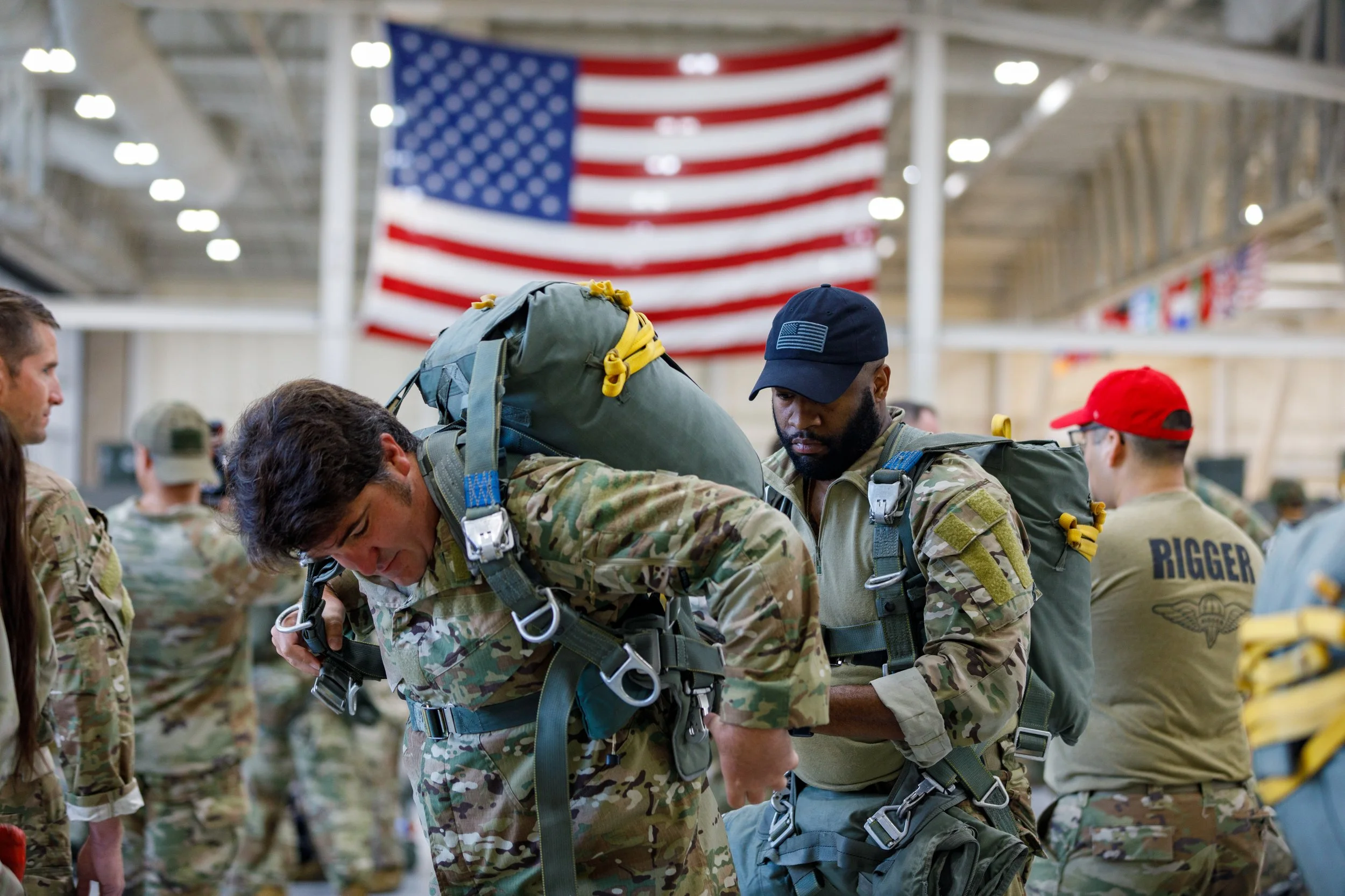 Two soldiers in camouflage uniforms preparing their gear inside a building with an American flag hanging in the background, surrounded by other soldiers.