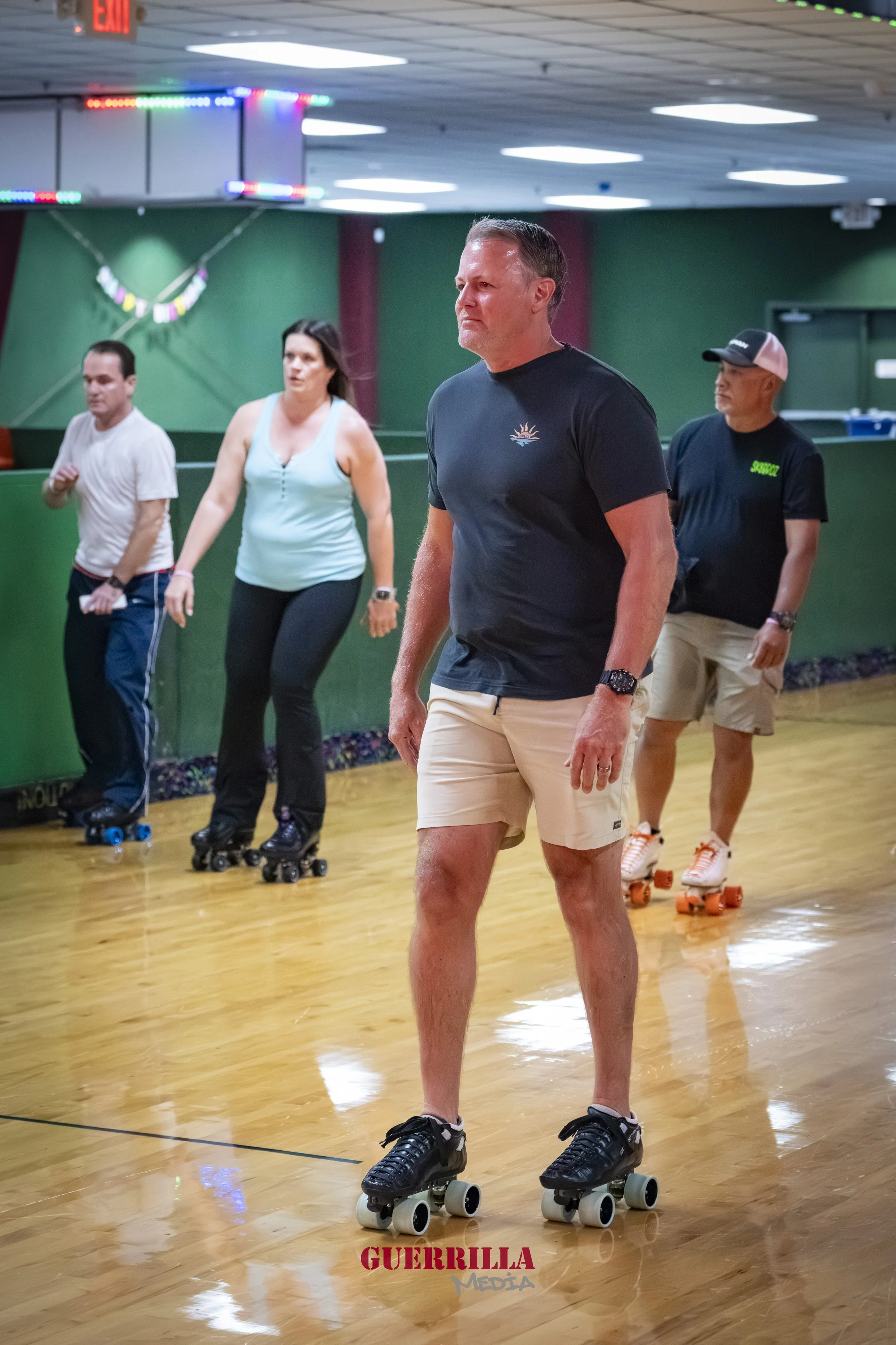 Four people roller skating in an indoor rink, the man in the front dressed in a black T-shirt and beige shorts, leading the group.