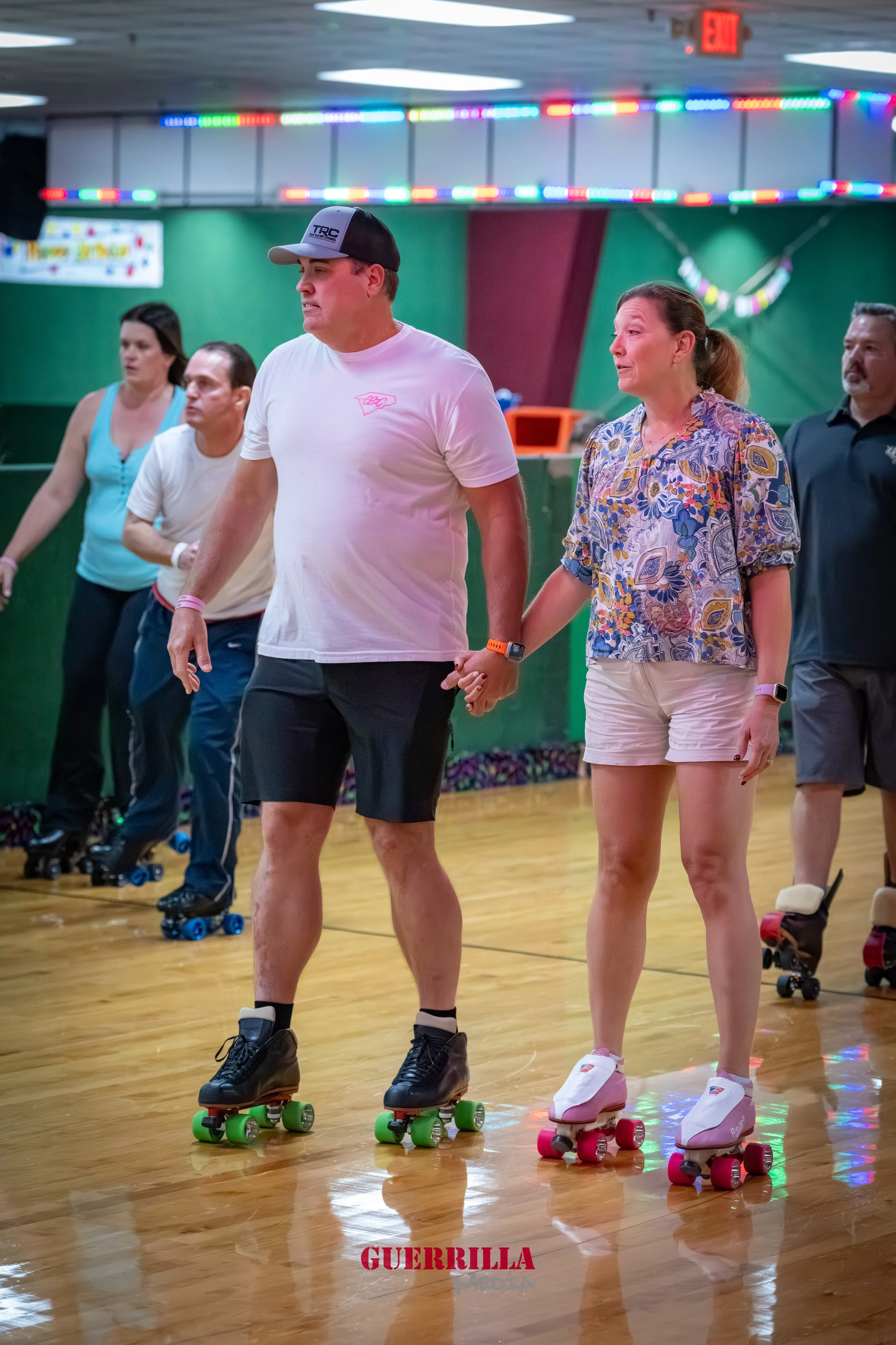 People roller skating holding hands in a roller rink with colorful lights and a green wall in the background.