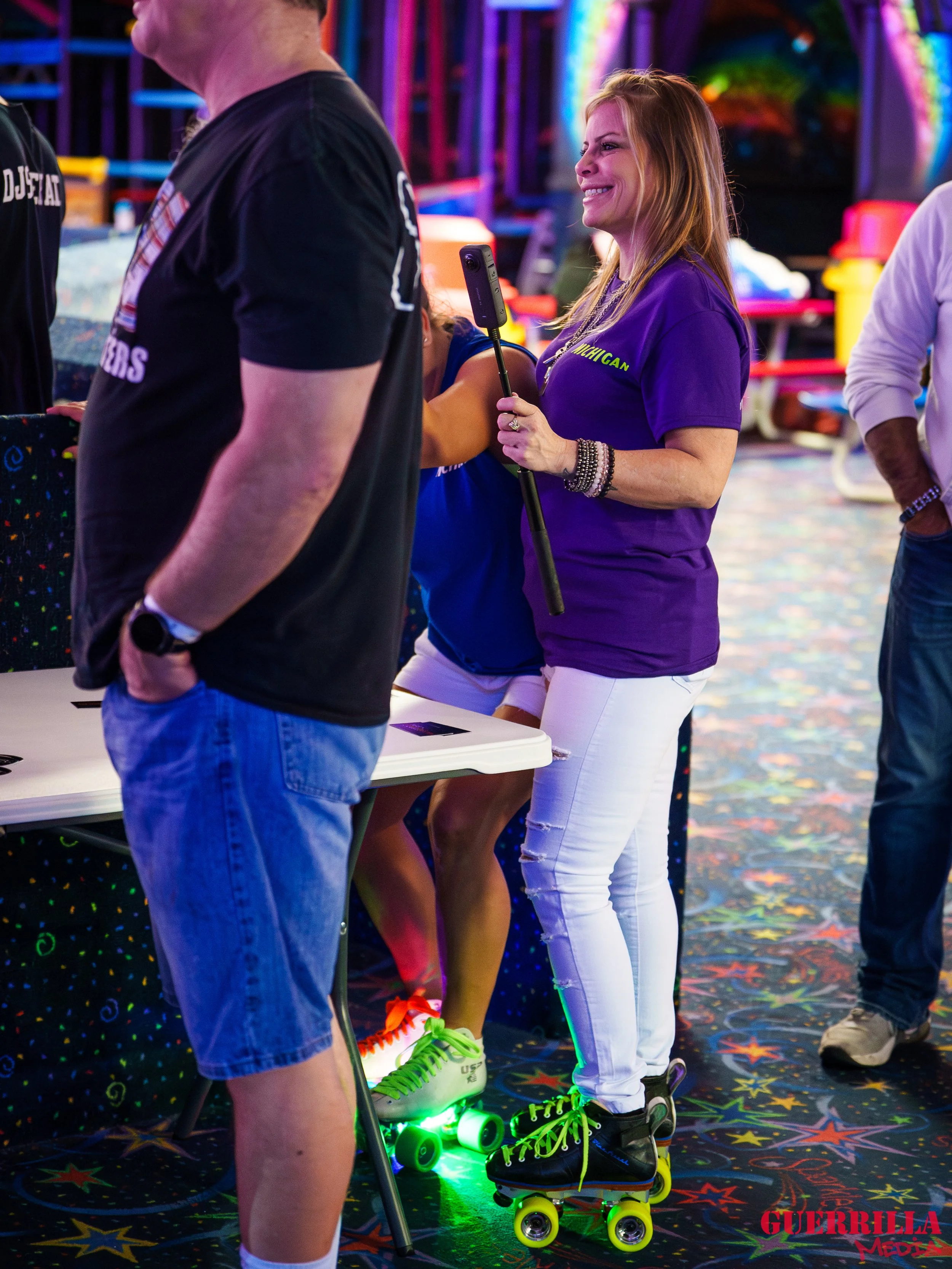 A woman wearing a purple shirt and white pants roller skating indoors at an amusement center, smiling and holding a selfie stick. The background features vibrant, colorful lighting and arcade or carnival games.
